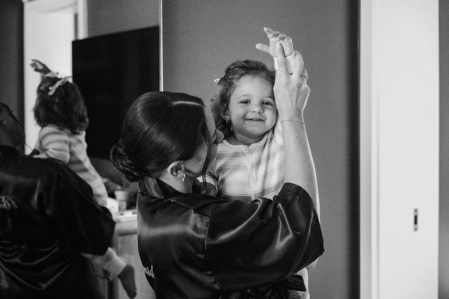 Black and white photo of a woman in satin robe lifting a smiling toddler girl during wedding preparations.