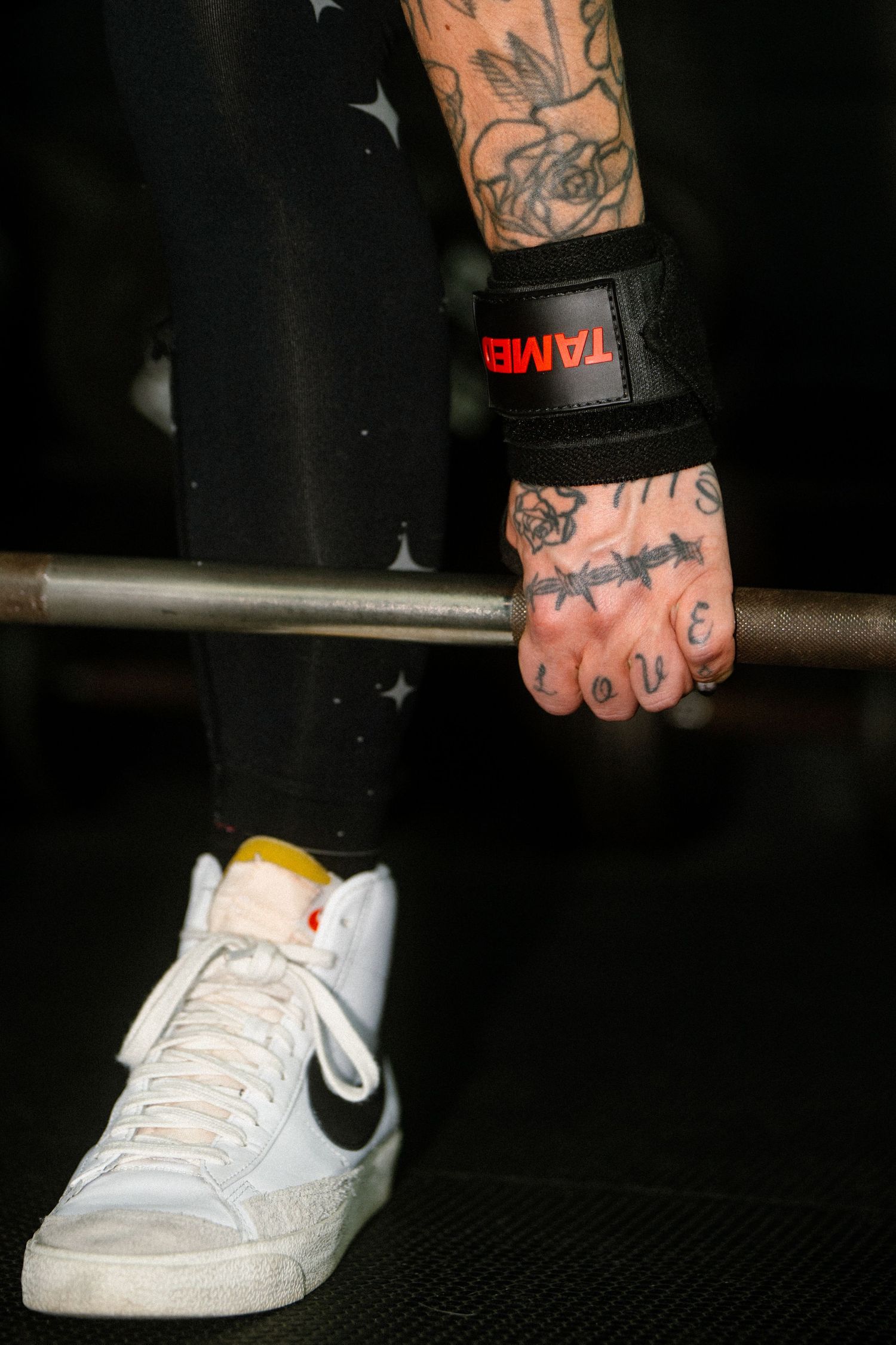 Tattooed hand gripping barbell with Tamer wrist wrap and white Nike sneaker visible below.
