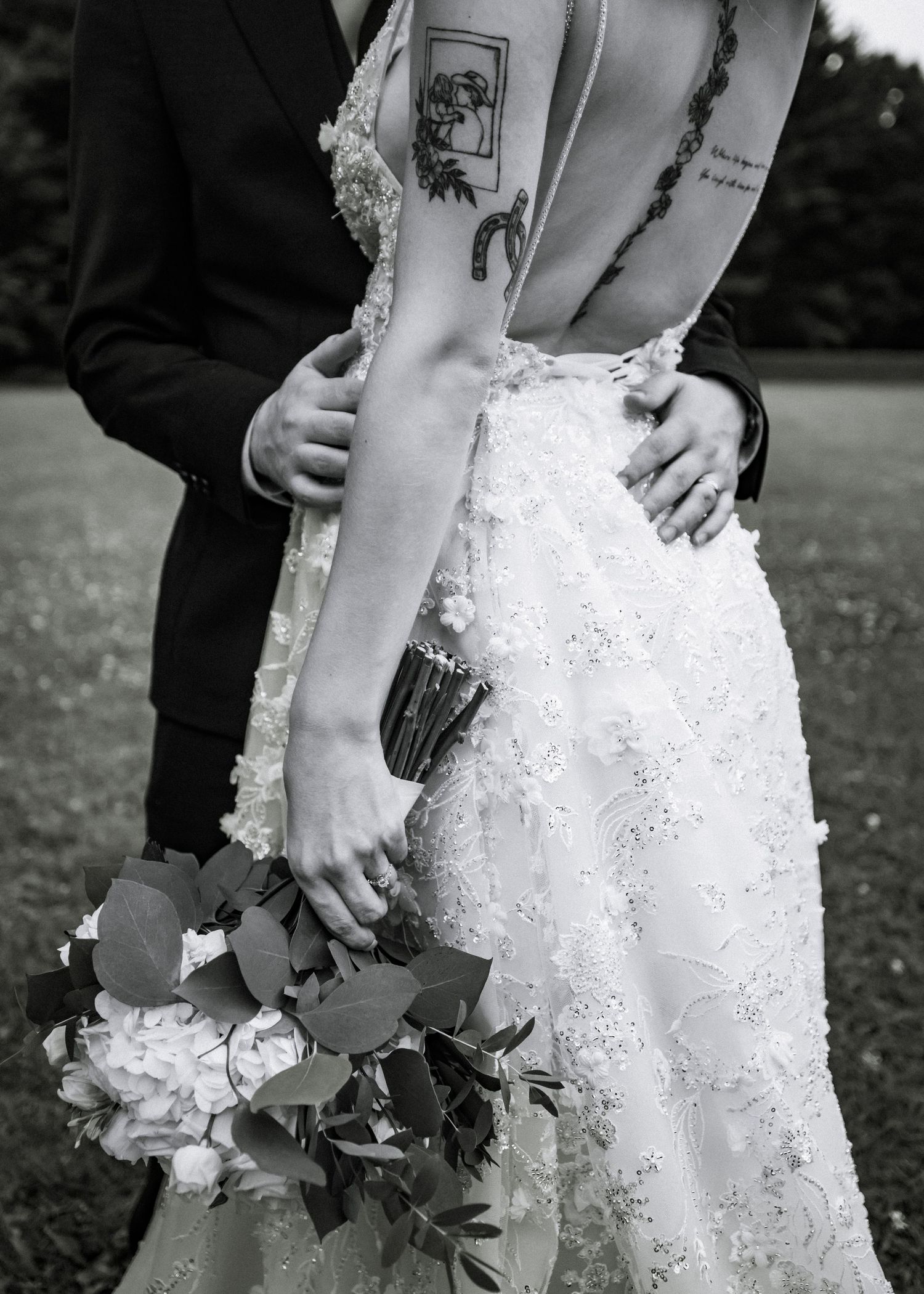 Black and white photo of bride and groom embracing, showing back tattoos and bridal bouquet.