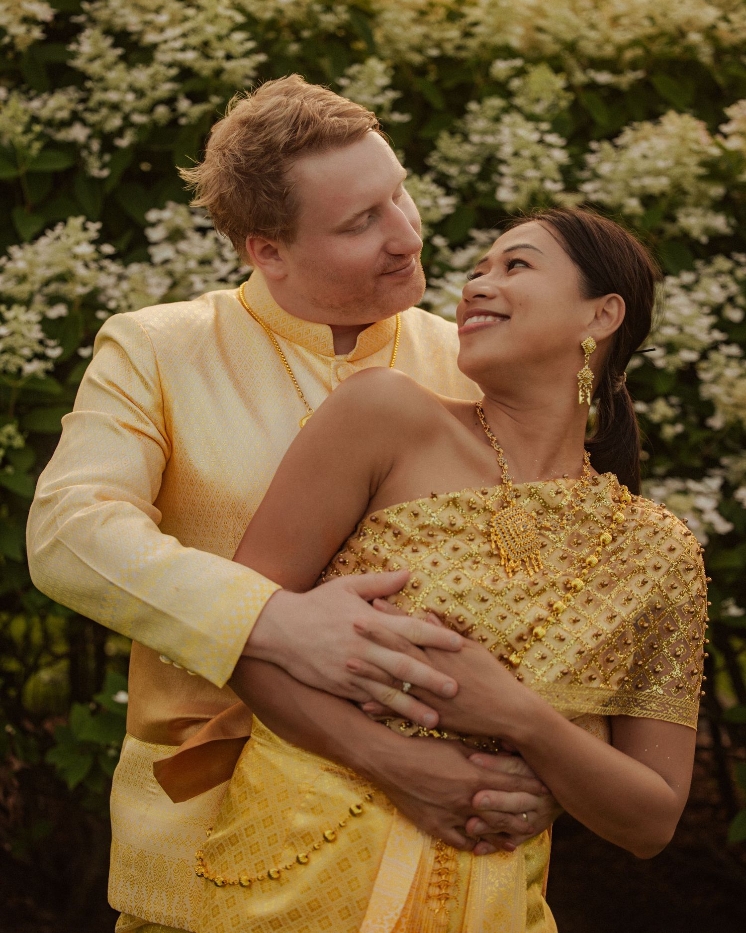 Couple embracing in matching golden traditional Thai wedding attire surrounded by white flowers outdoors.