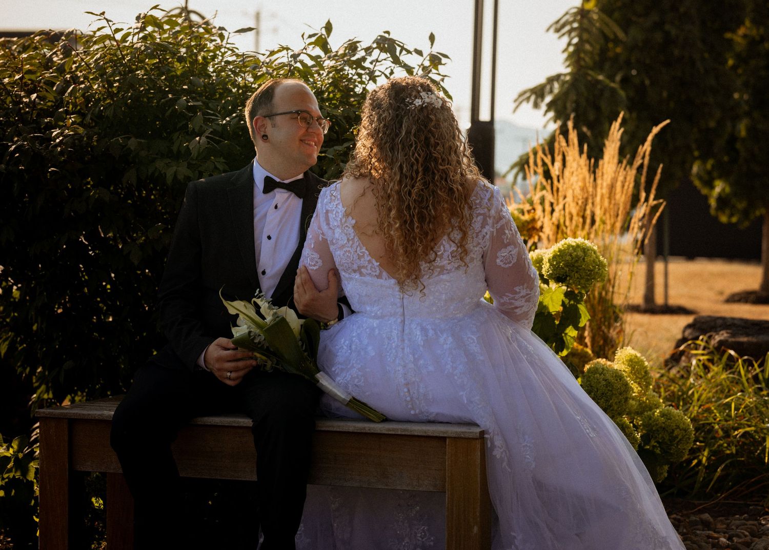 Bride in white lace gown and groom in black tuxedo sit together outdoors, sharing a romantic moment at their wedding.