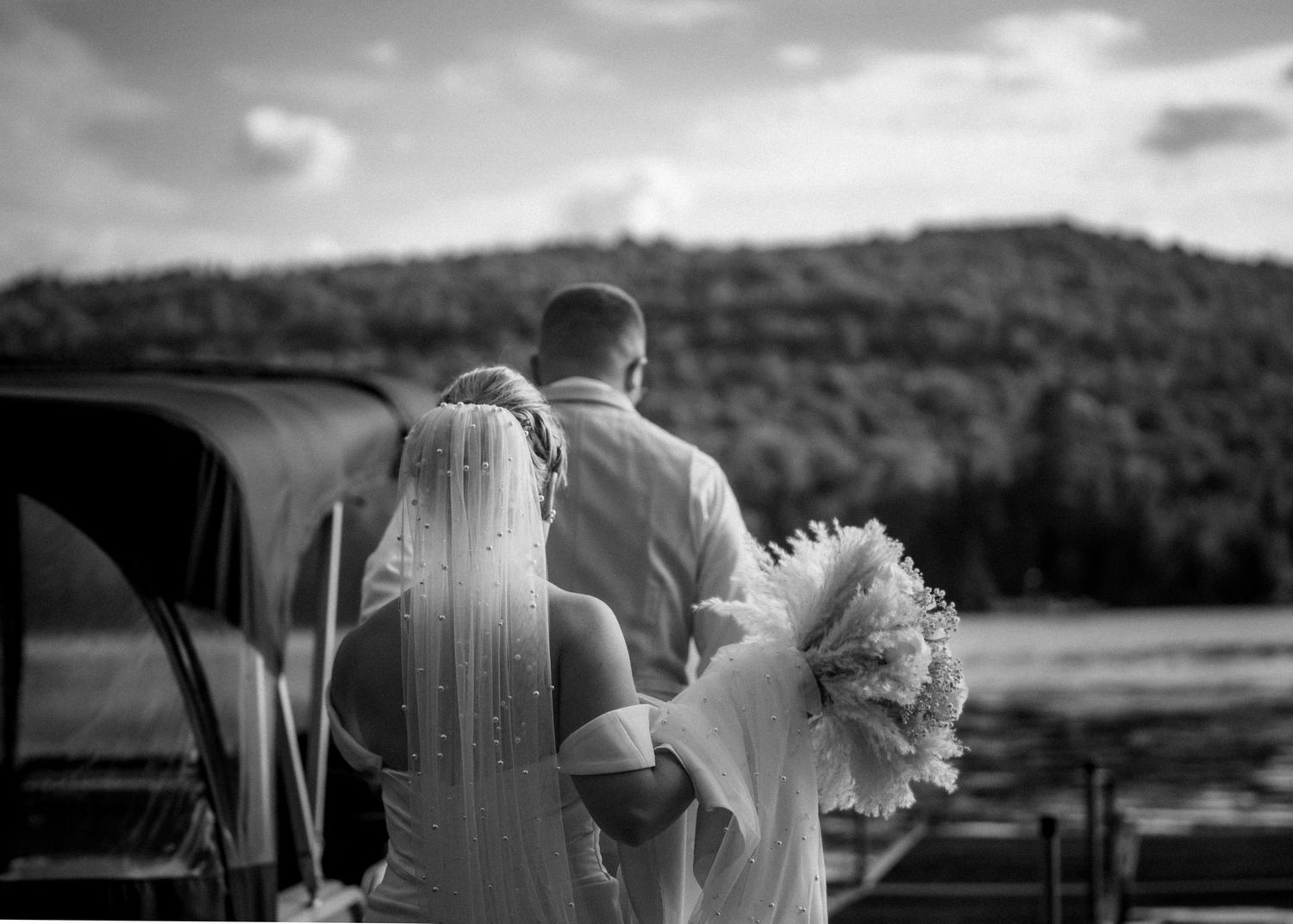 Black and white photo of a bride and groom walking hand in hand toward a lake with forested hills in the background.