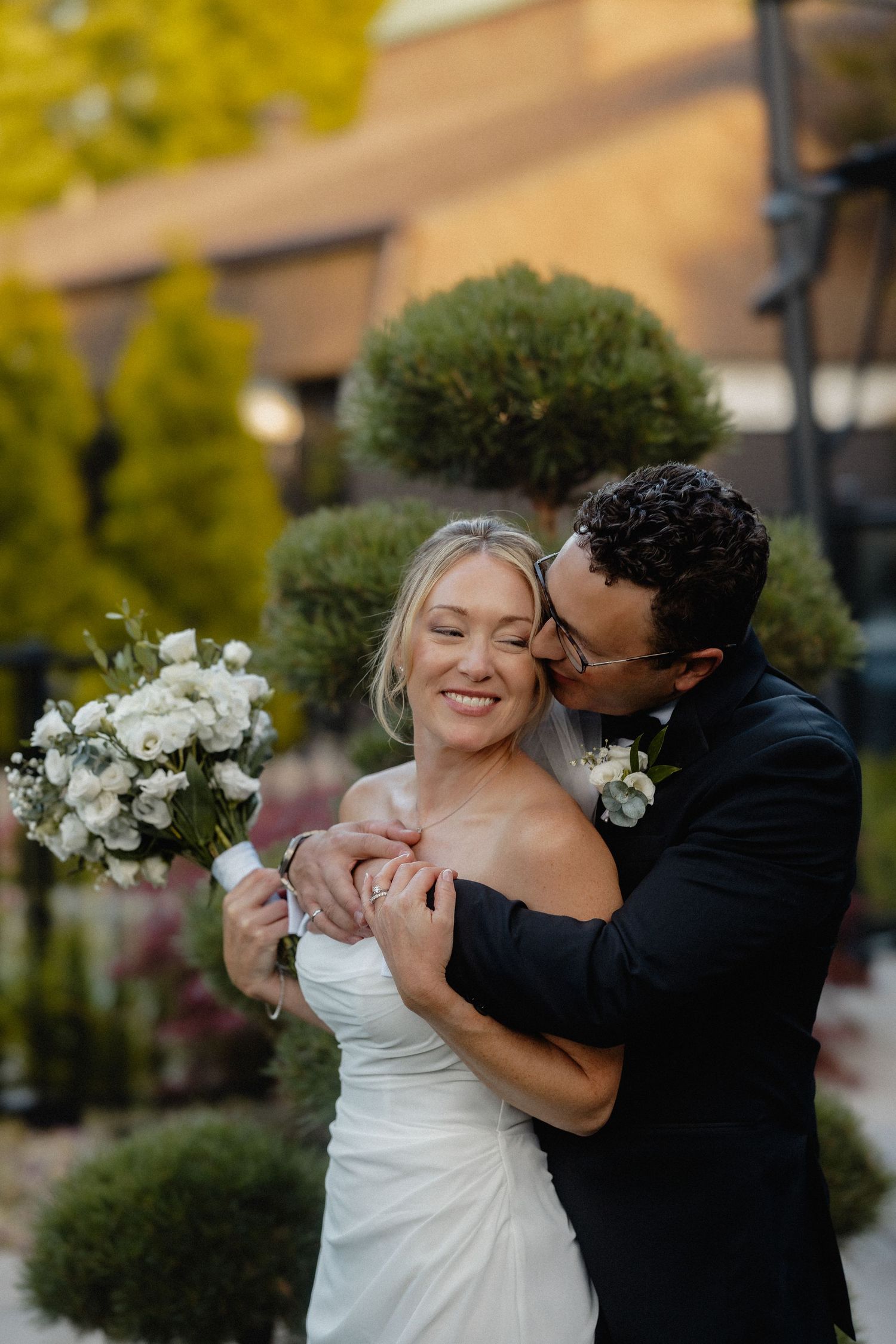 Groom in black suit kisses bride on cheek as she smiles holding white floral bouquet outdoors.