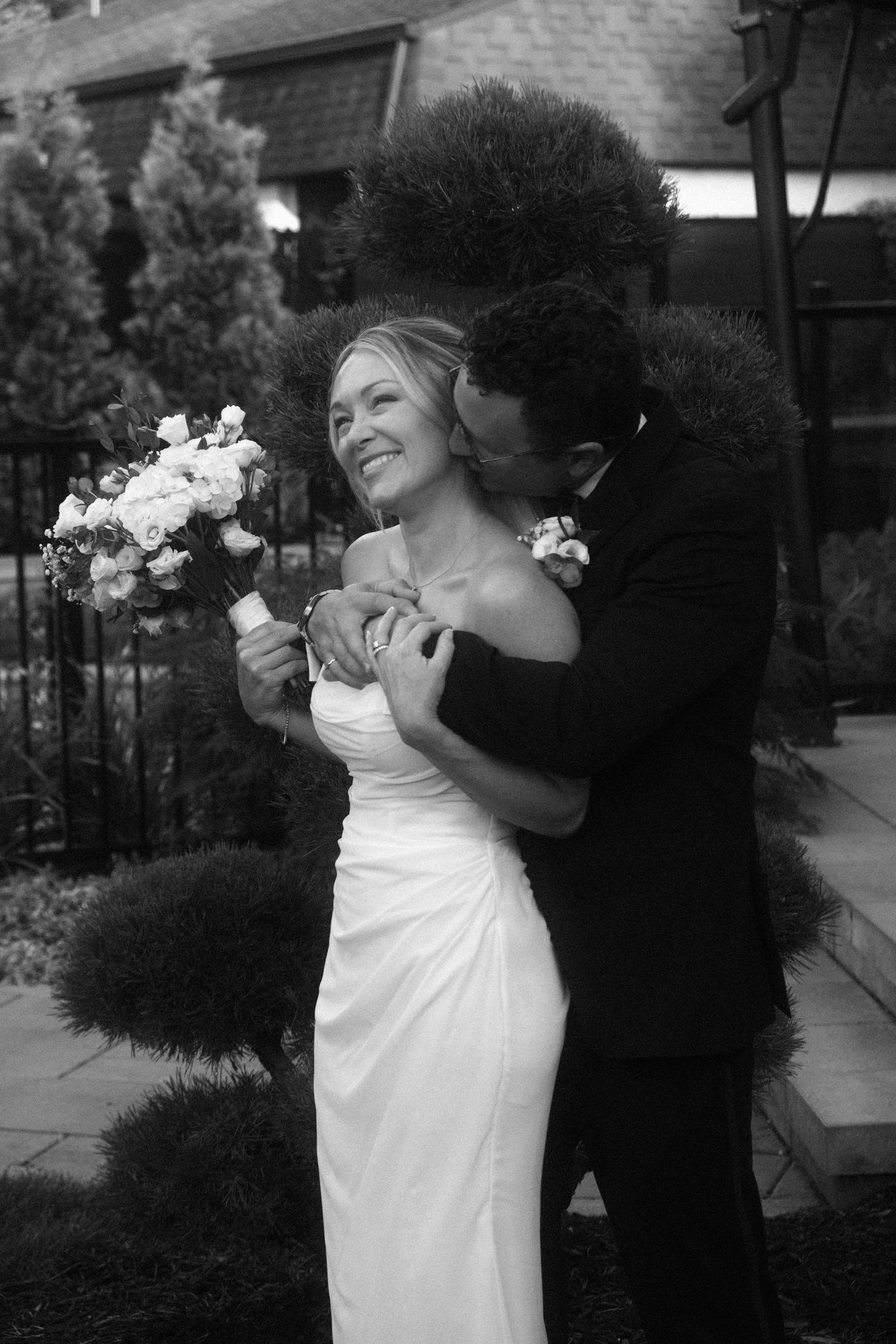 Elegant black and white wedding portrait of bride smiling with bouquet as groom tenderly kisses her cheek.