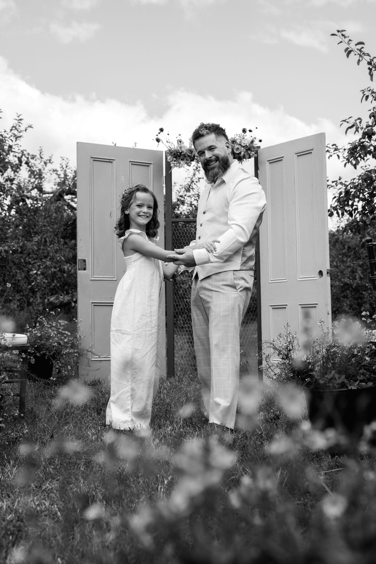 Black and white photo of bearded groom holding hands with flower girl by decorative doors.