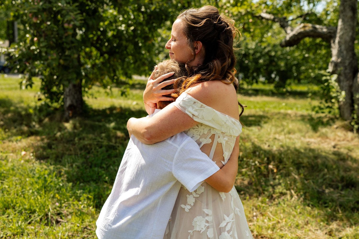 Bride in lace off-shoulder gown embracing groom outdoors in a sunlit garden setting.