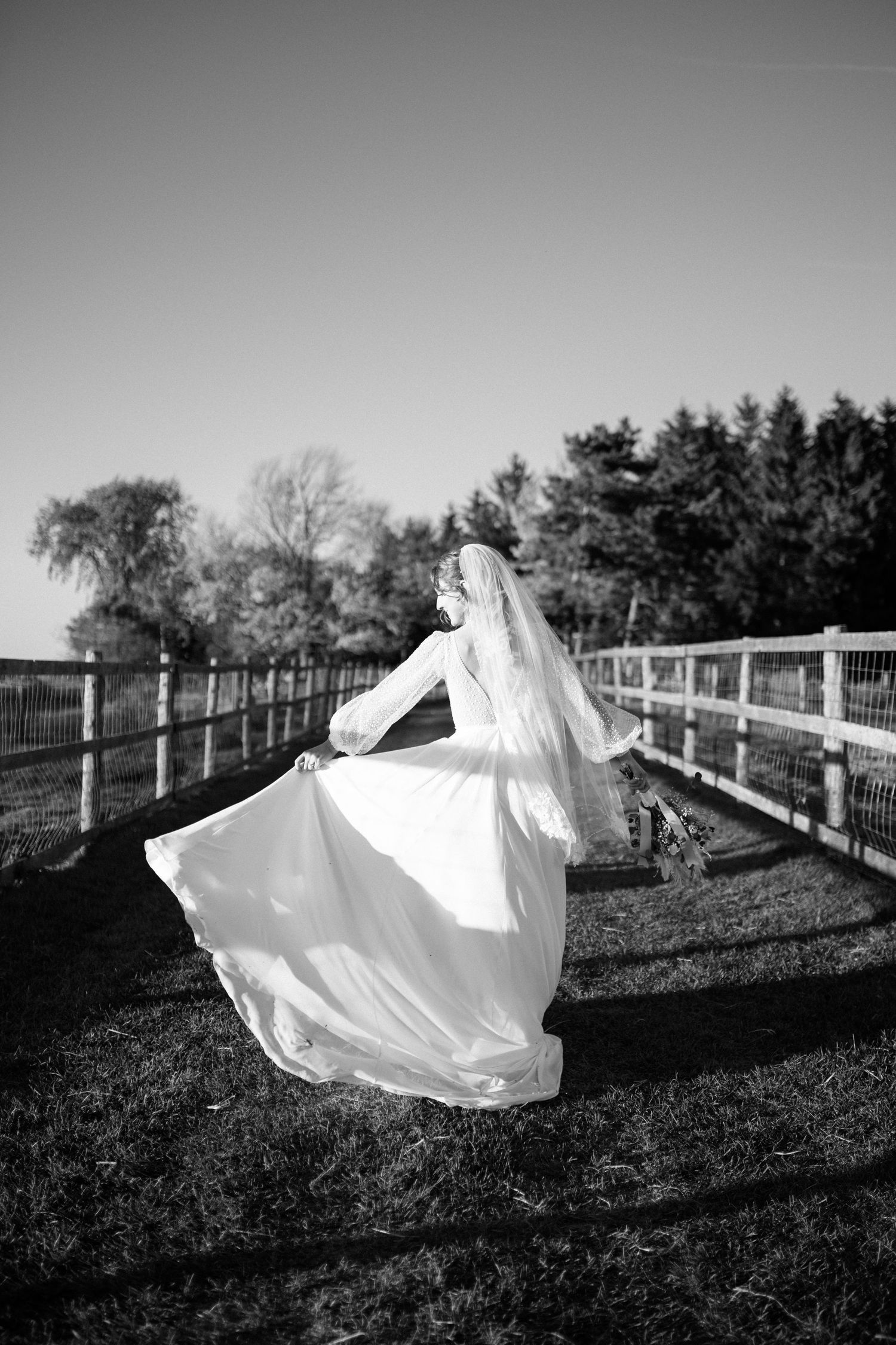 Black and white photo of bride twirling in white gown by a fence in an outdoor countryside setting.