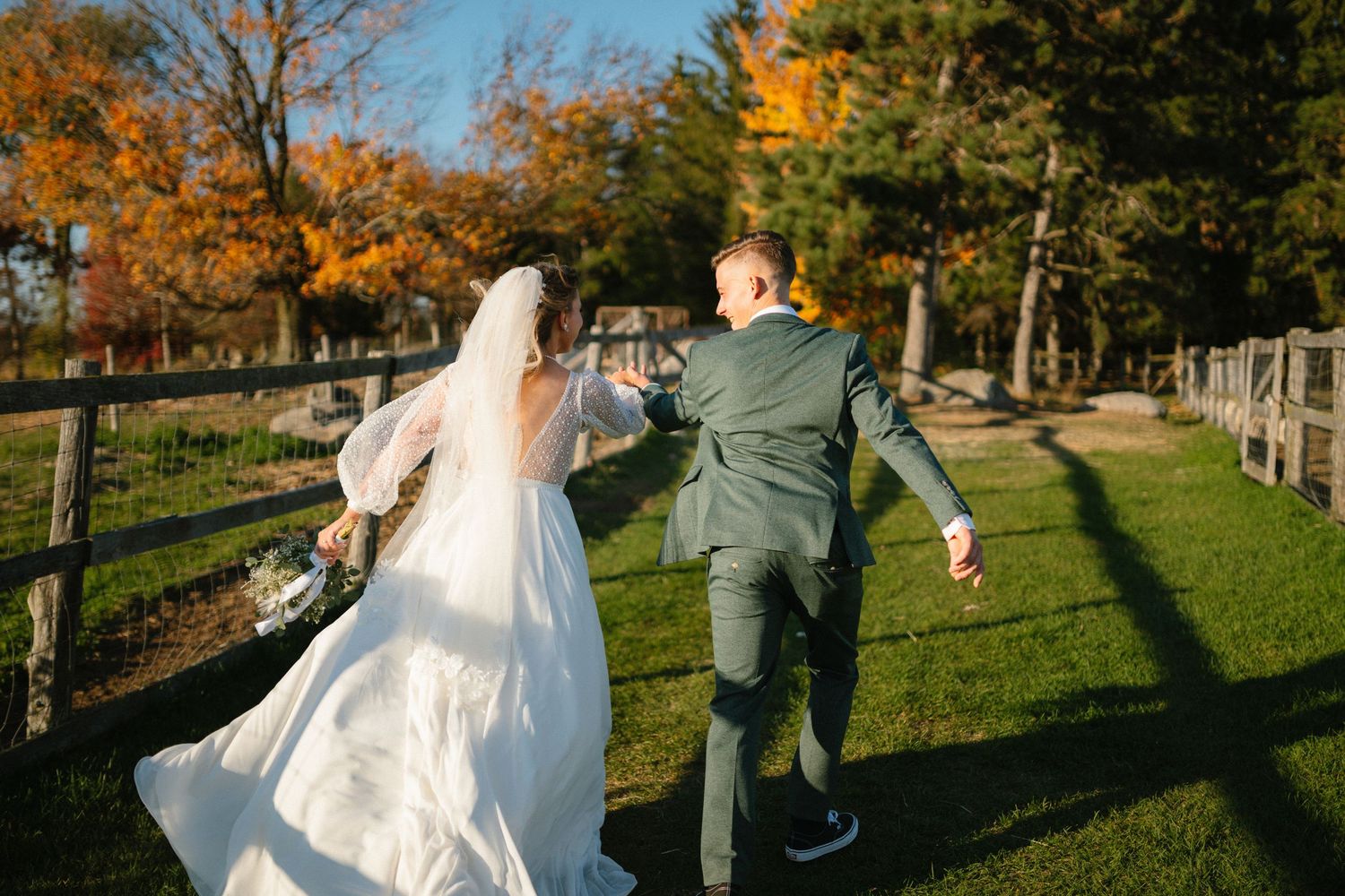 A bride and groom joyfully run hand in hand through a sunny green field with autumn trees.