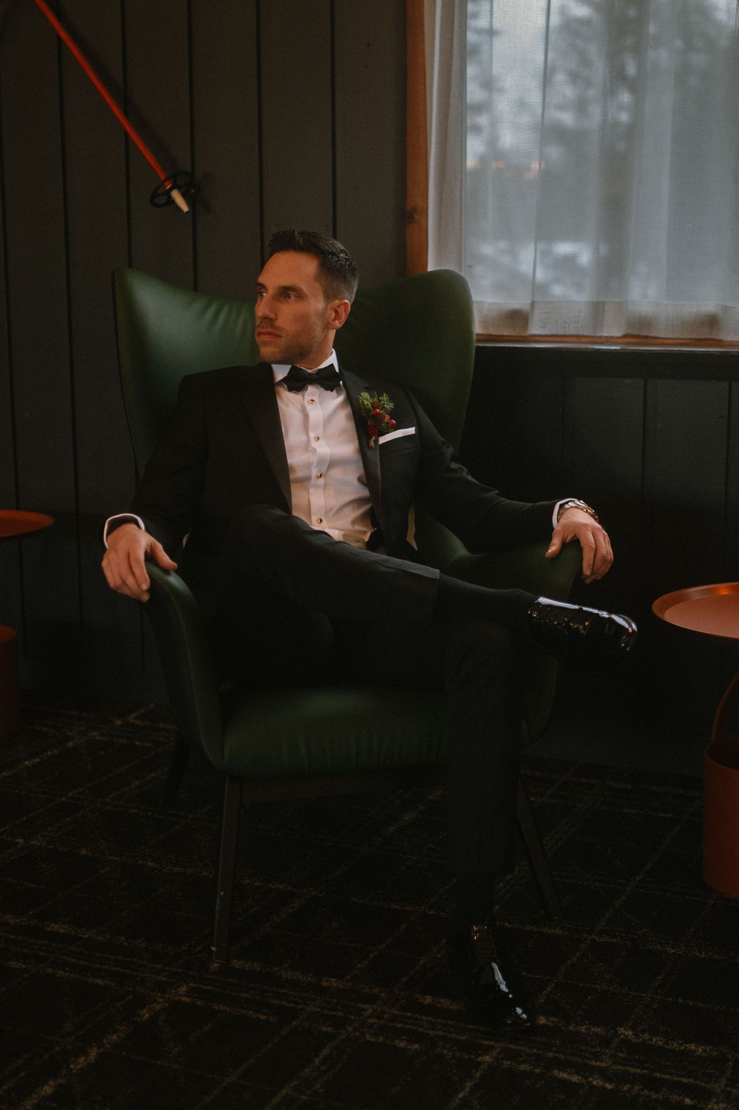 A groom in a black tuxedo with bow tie sits elegantly in a green velvet chair, gazing thoughtfully to the side.