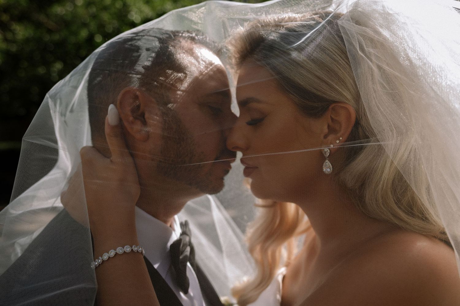 Bride and groom sharing intimate kiss under flowing white veil in romantic outdoor portrait.