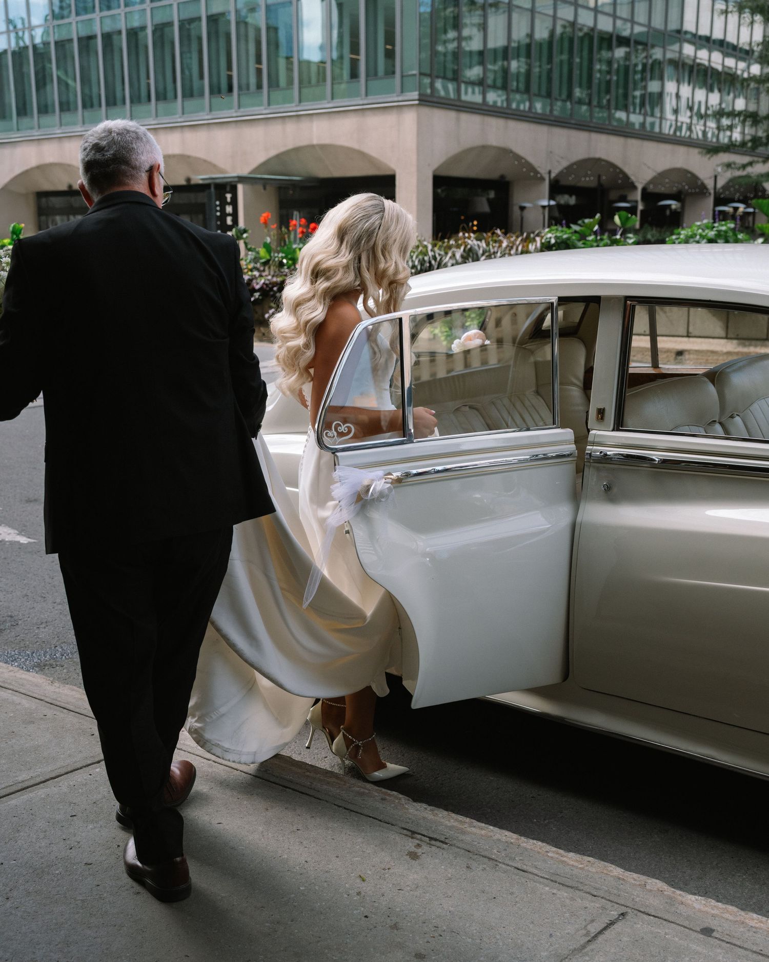 Bride in backless wedding gown entering a vintage white car with escort on wedding day.