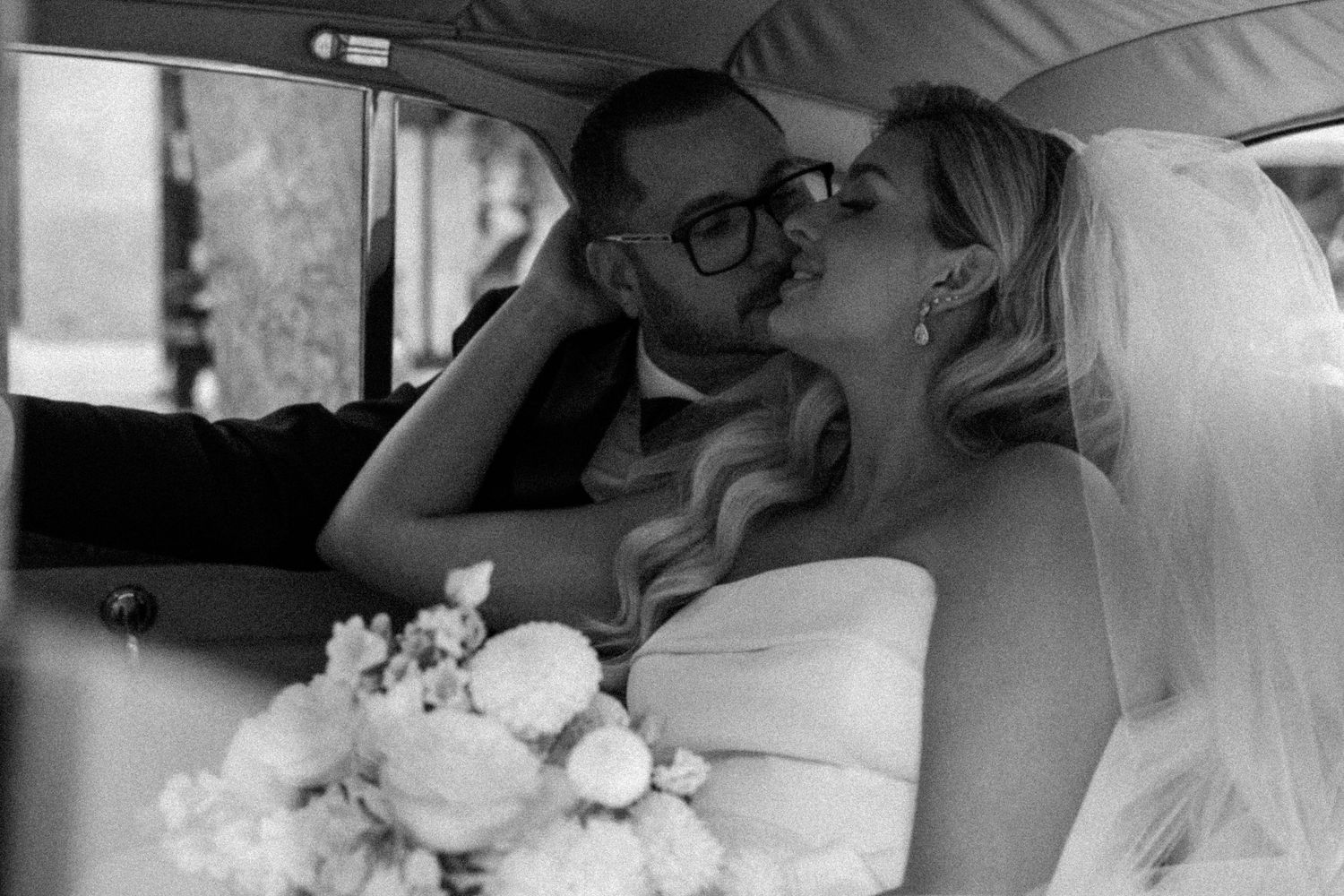 Newlyweds sharing a romantic kiss inside a vintage car after their wedding ceremony.