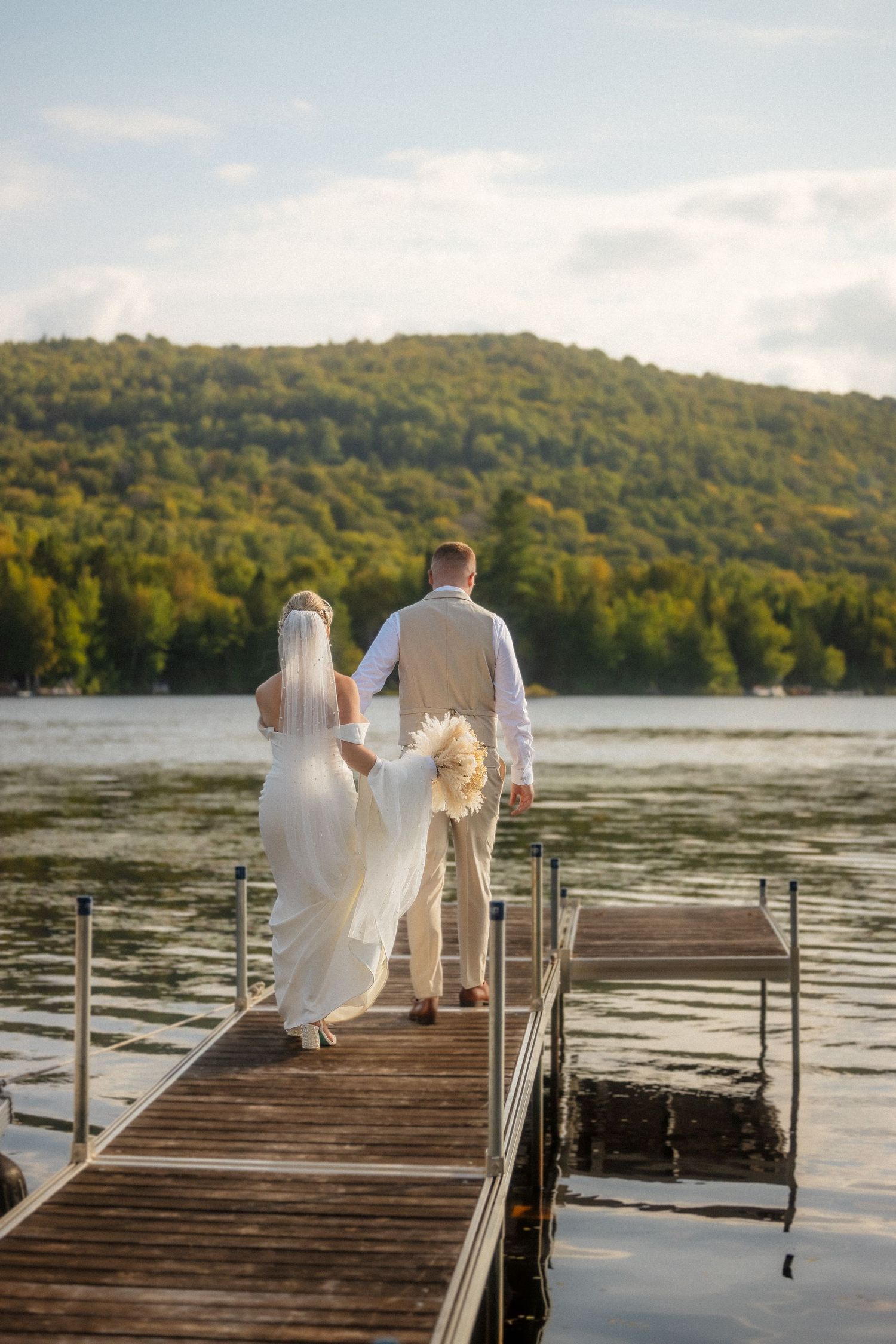 Bride and groom walking hand in hand down a wooden dock toward a serene lake surrounded by lush green hills.