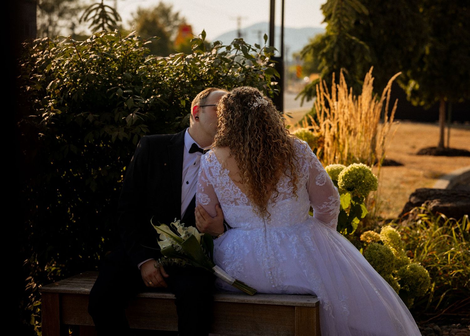 Bride and groom sharing a romantic kiss while seated outdoors in golden hour light, surrounded by lush greenery.