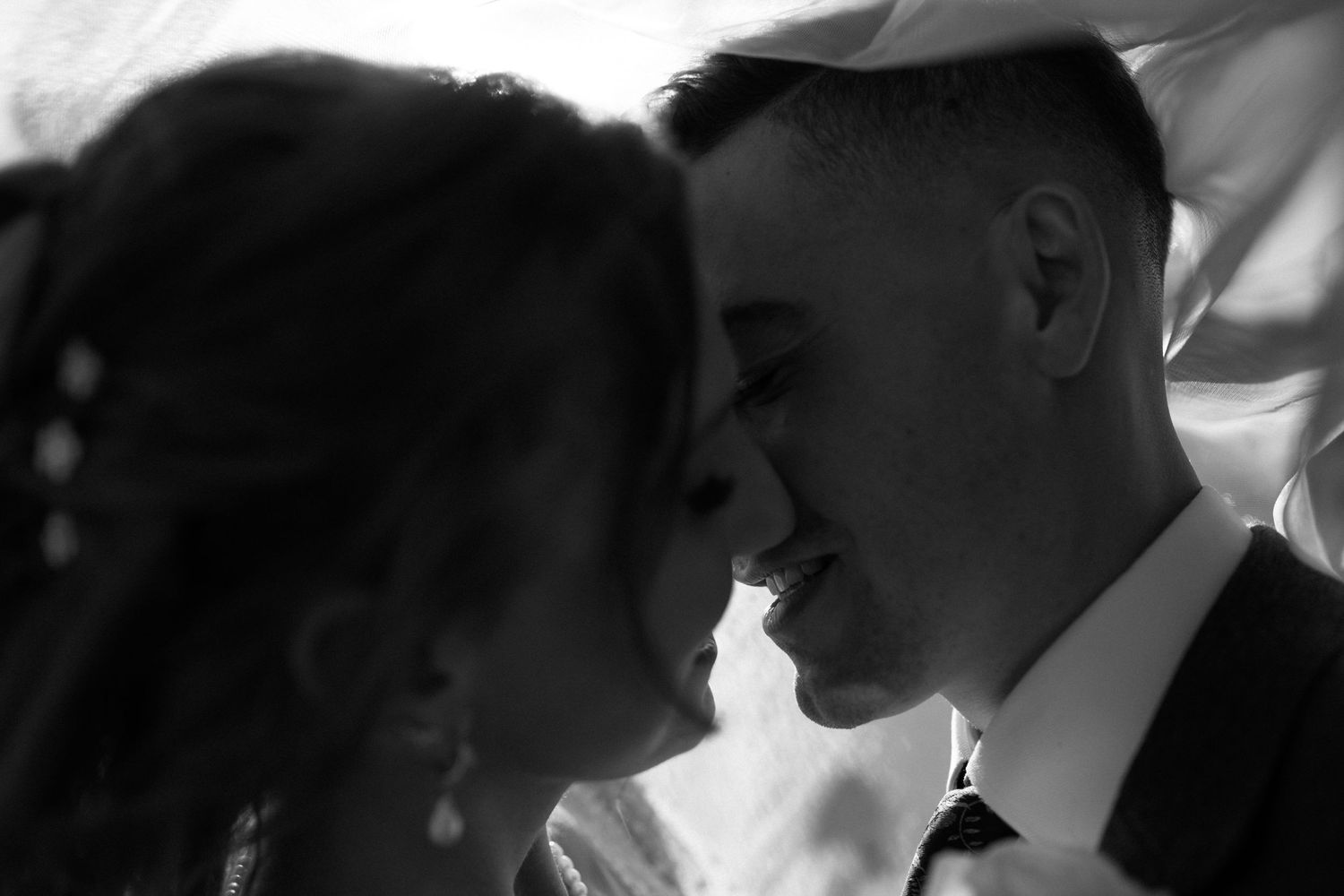 Romantic black and white close-up of a smiling bride and groom nearly kissing under a veil.