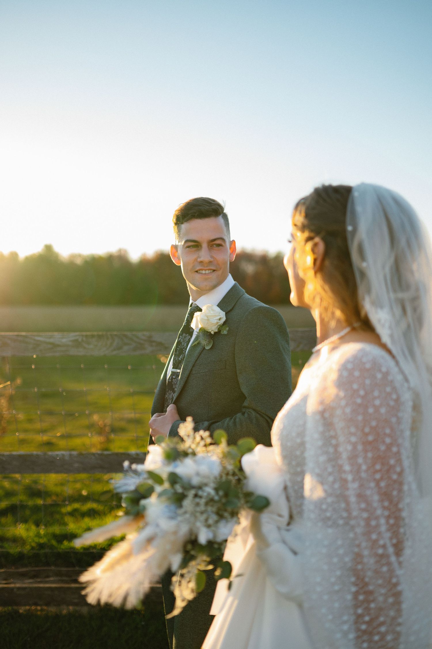 A smiling groom in a grey suit looks at his bride holding a white bouquet during a golden hour farm wedding.