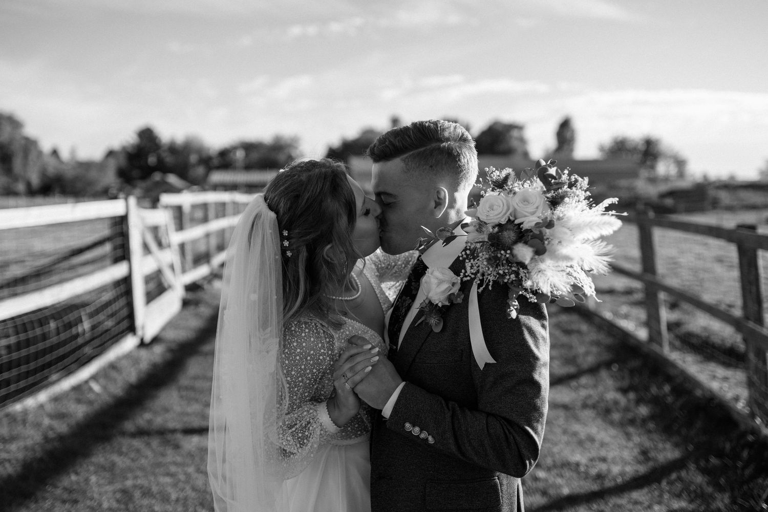 Black and white portrait of bride and groom touching foreheads by white fence outdoors.