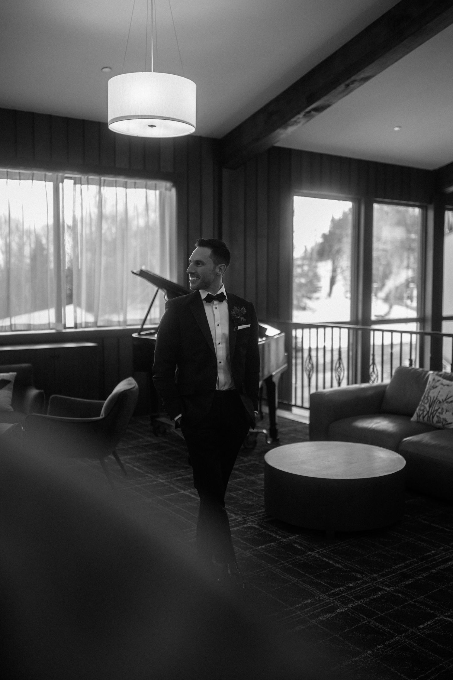 Dapper groom in black tuxedo standing in a modern lodge with snowy mountain views through large windows.