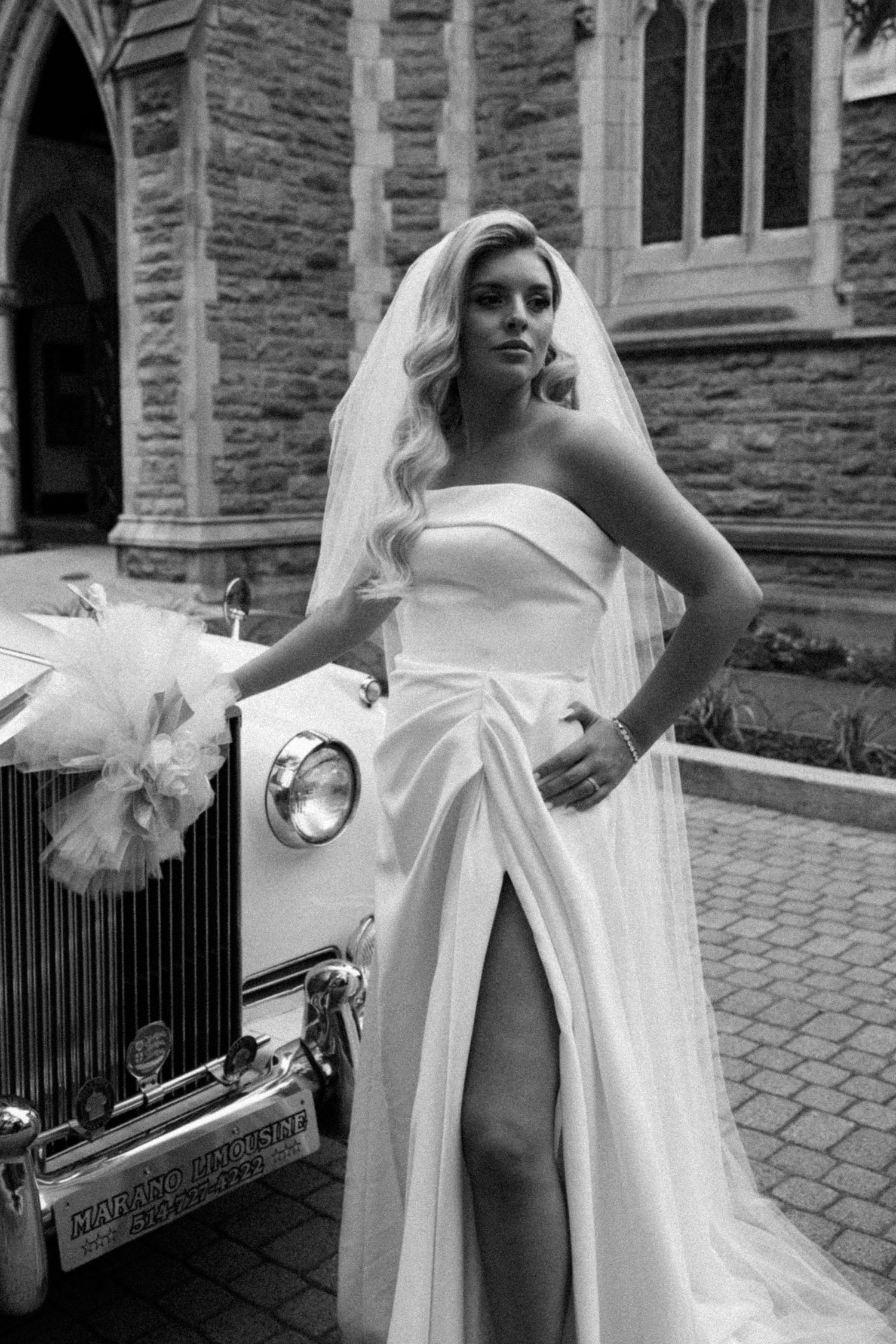 Elegant bride in strapless white gown with thigh slit and veil posing beside vintage Rolls-Royce outside stone church.