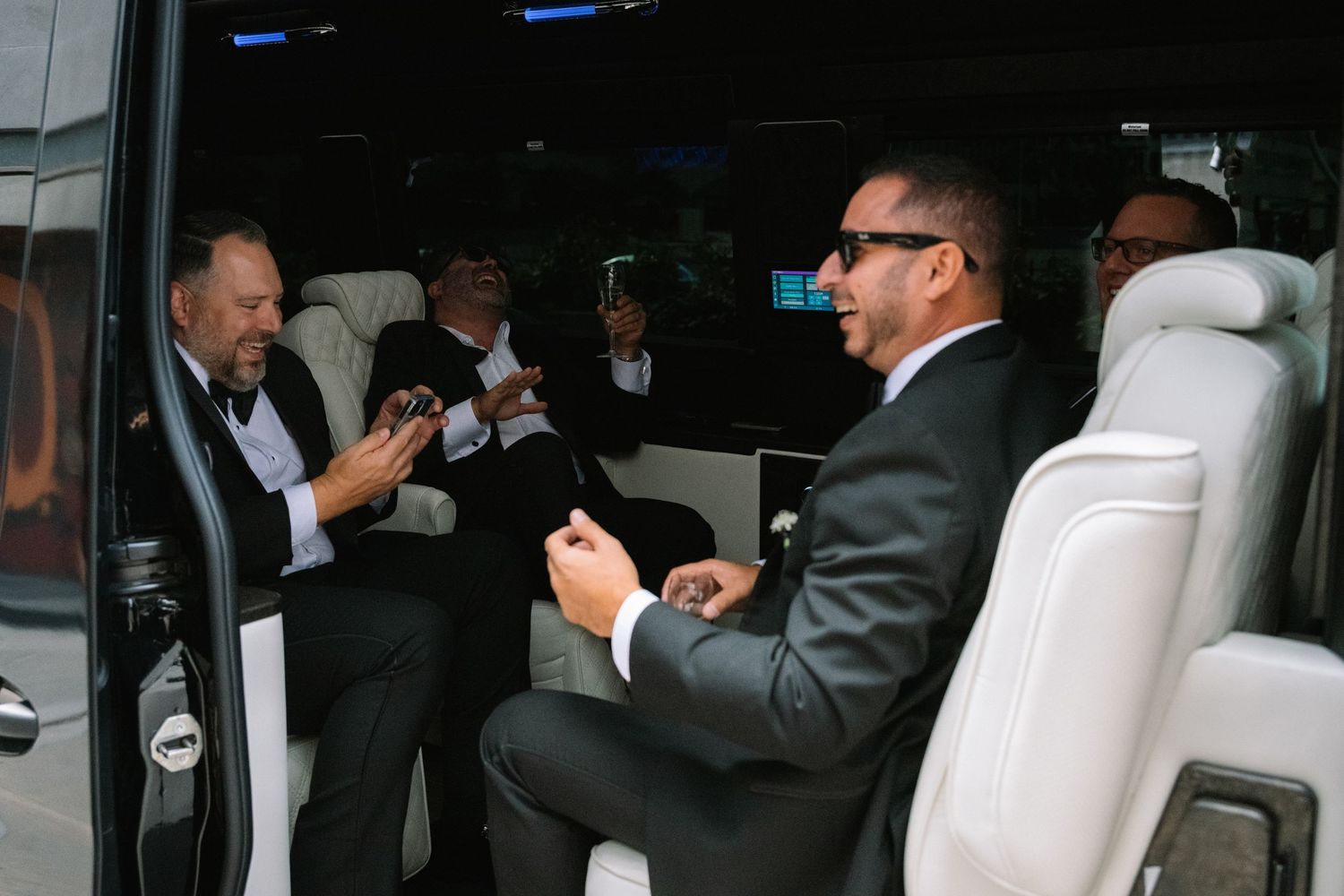 Groomsmen in black tuxedos laughing and celebrating inside luxury white party bus before wedding.