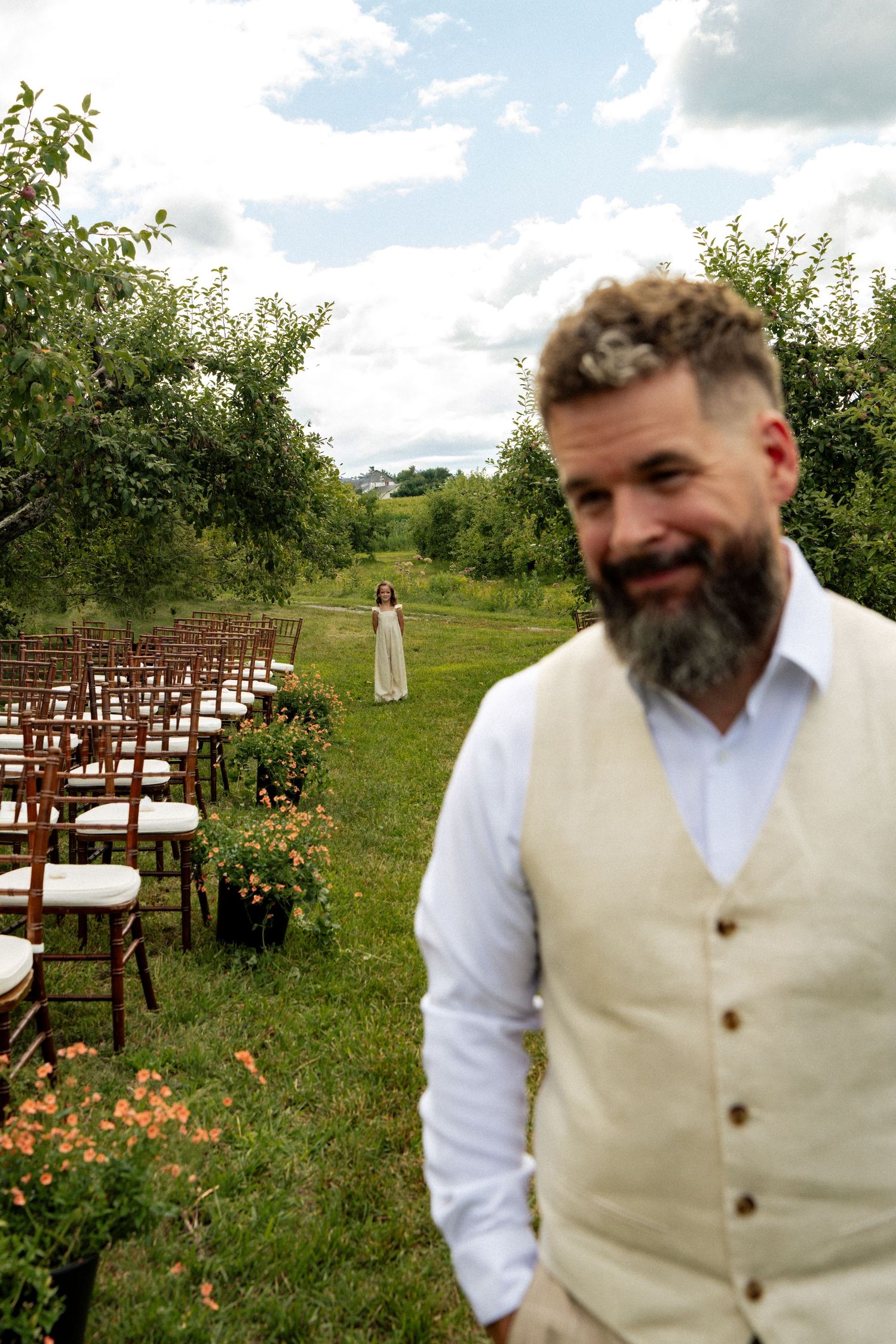 Bearded groom in linen vest smiles at outdoor wedding venue with rows of wooden chairs in orchard.