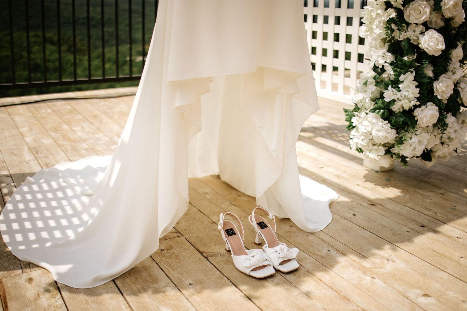Elegant white bridal heels placed on wooden deck beside flowing wedding gown near floral arrangement.