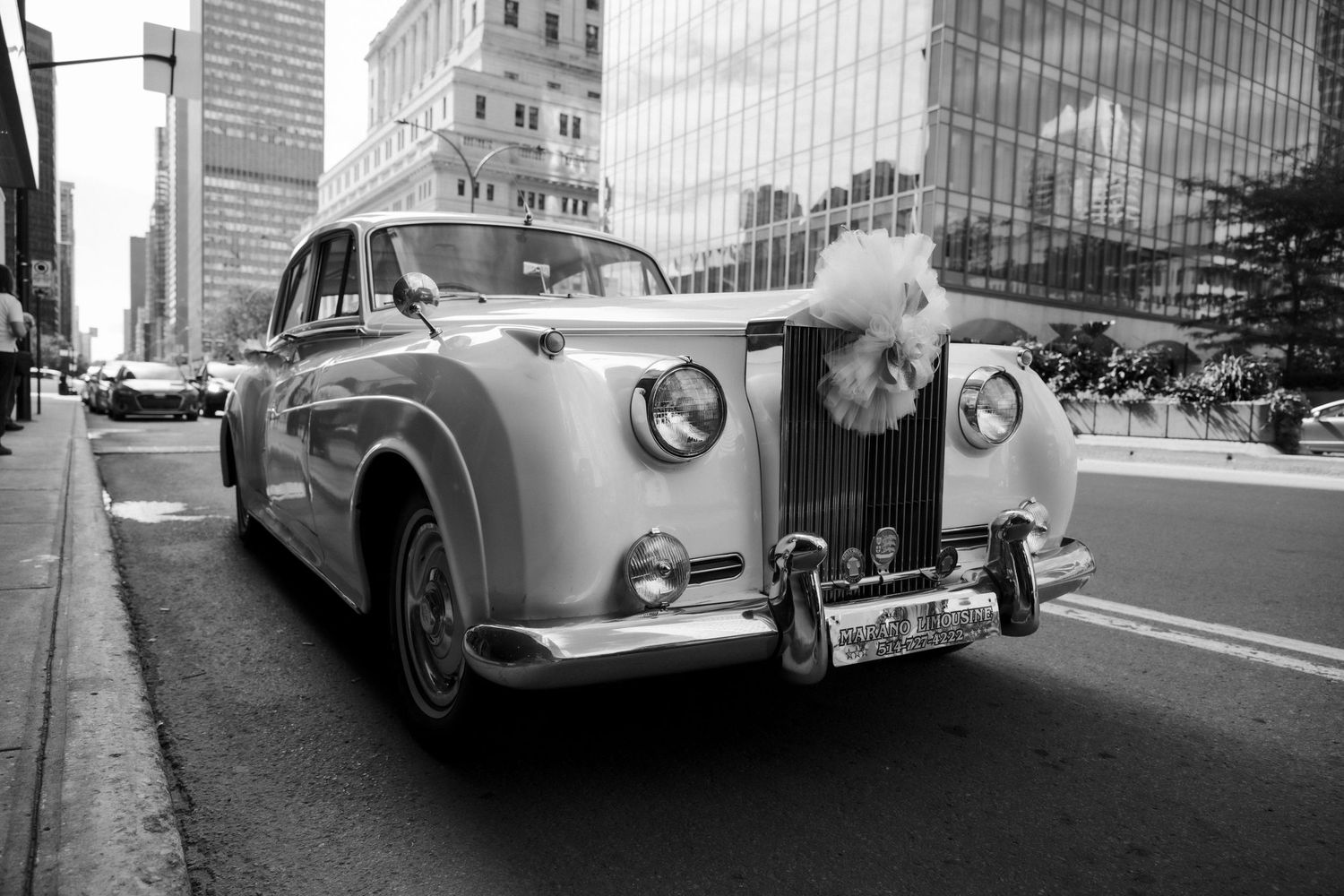 Vintage Rolls-Royce wedding car decorated with white flowers driving through city streets in black and white.