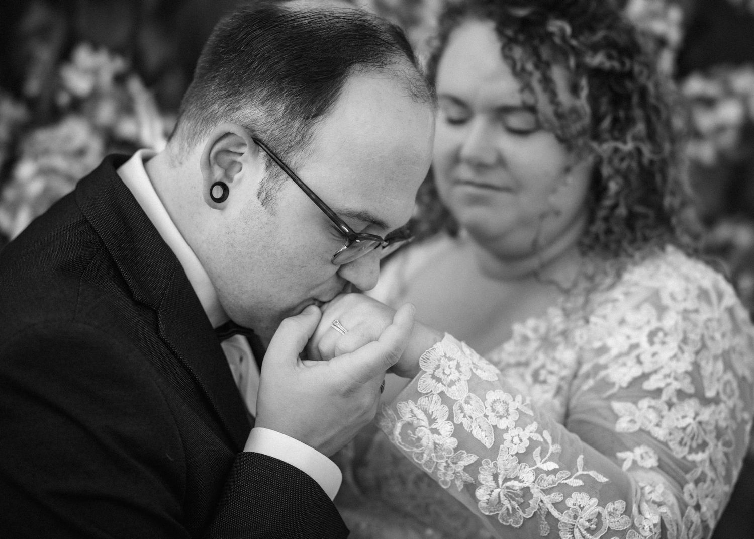 Groom in black suit kissing bride's hand during wedding ceremony, bride in lace dress smiling, black and white photo.