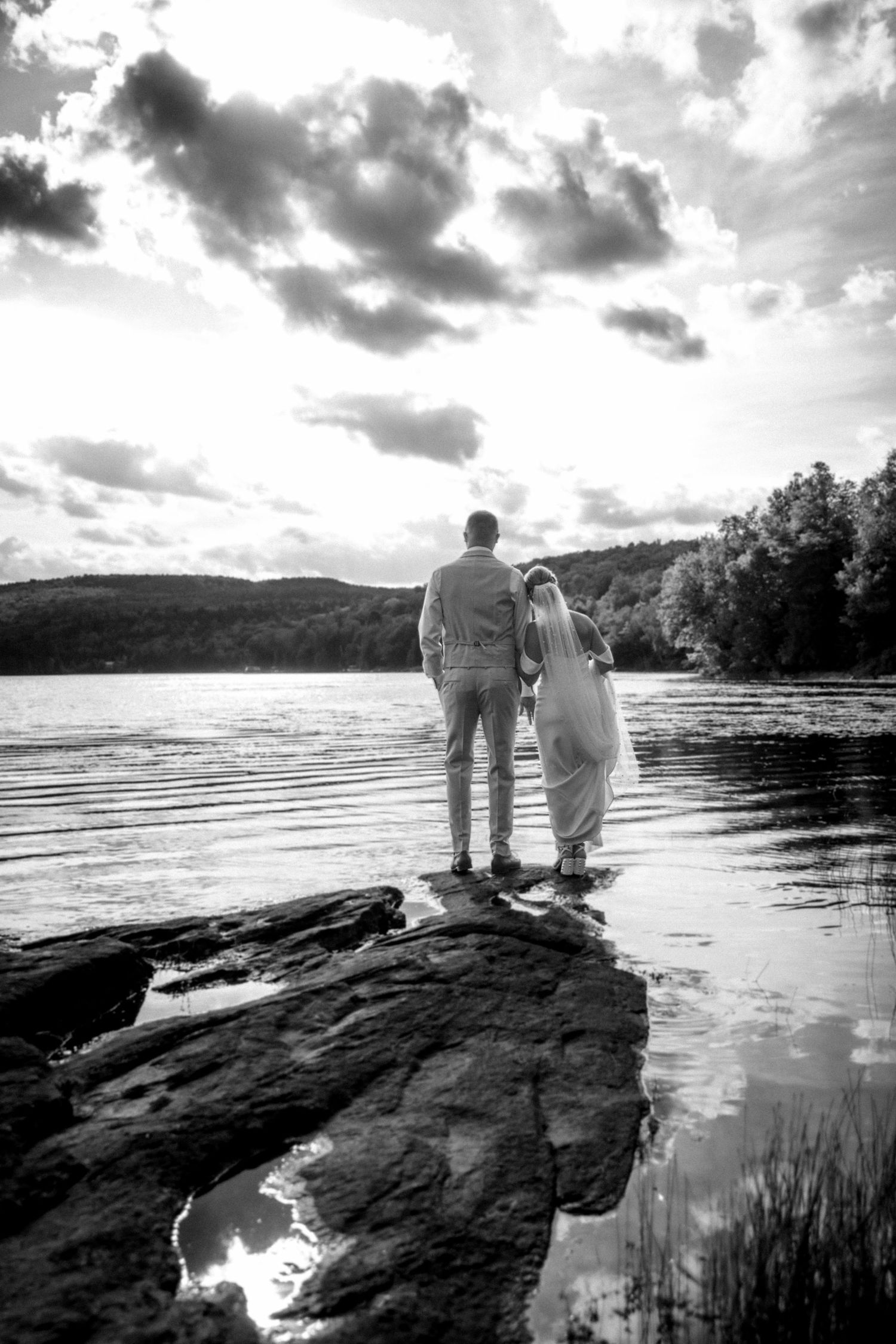 Black and white photo of a bride and groom standing on rocks by a tranquil lake on their wedding day.