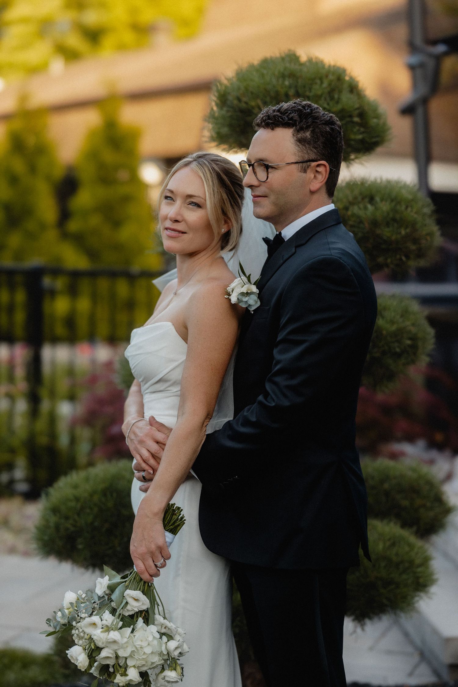 Bride and groom pose outdoors in formal wedding attire, she holds white bouquet, surrounded by manicured garden greenery.
