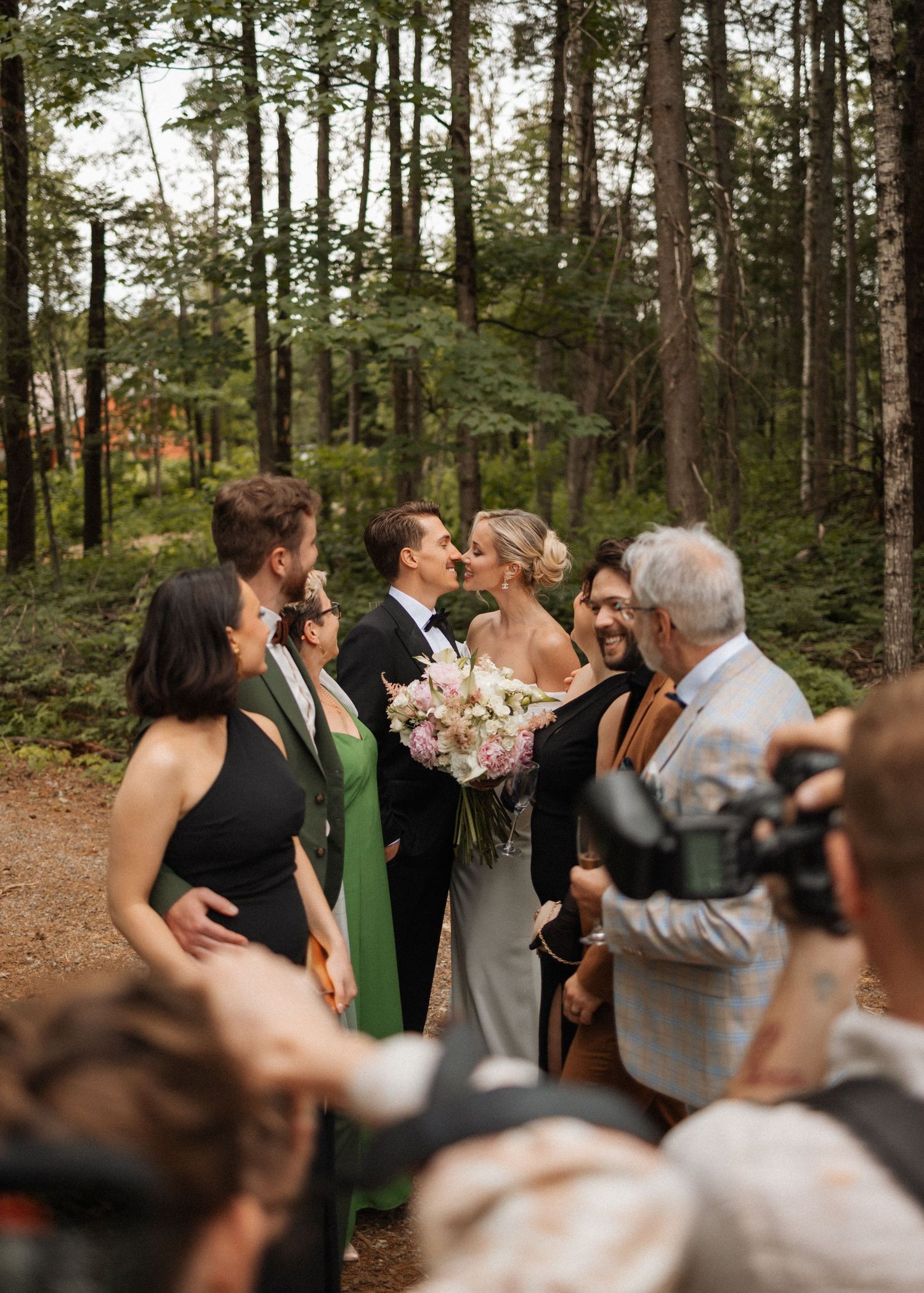 Bride and groom share a kiss surrounded by wedding guests in a lush forest setting, bride holding a pink bouquet.