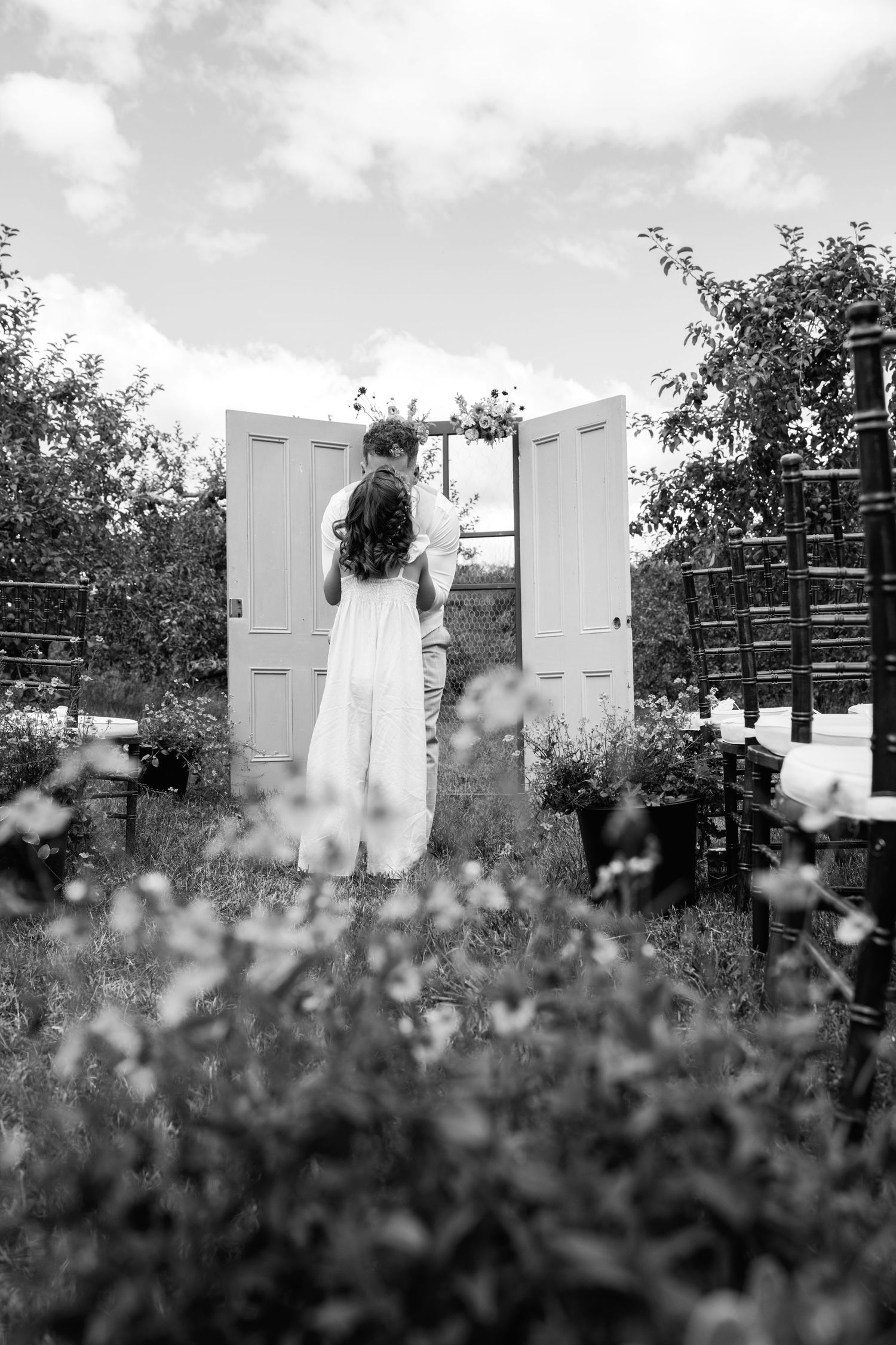 Black and white photo of a bride and groom embracing before open white doors at an outdoor wedding.
