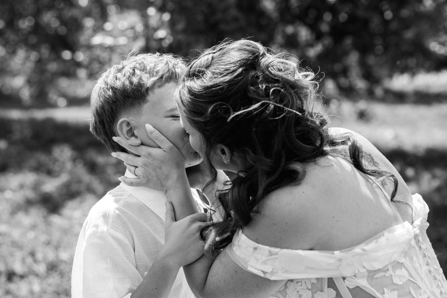 Black and white photo of a bride in off-shoulder gown receiving a kiss on the cheek from a young boy outdoors.