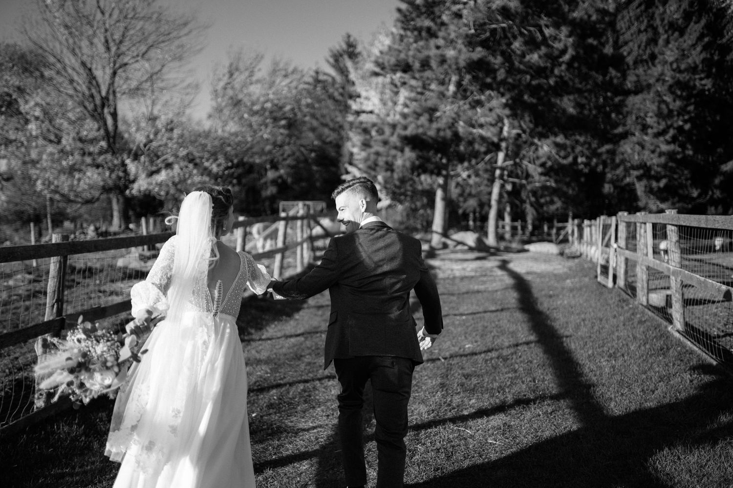 Black and white photo of bride and groom walking hand in hand along a fenced path outdoors.