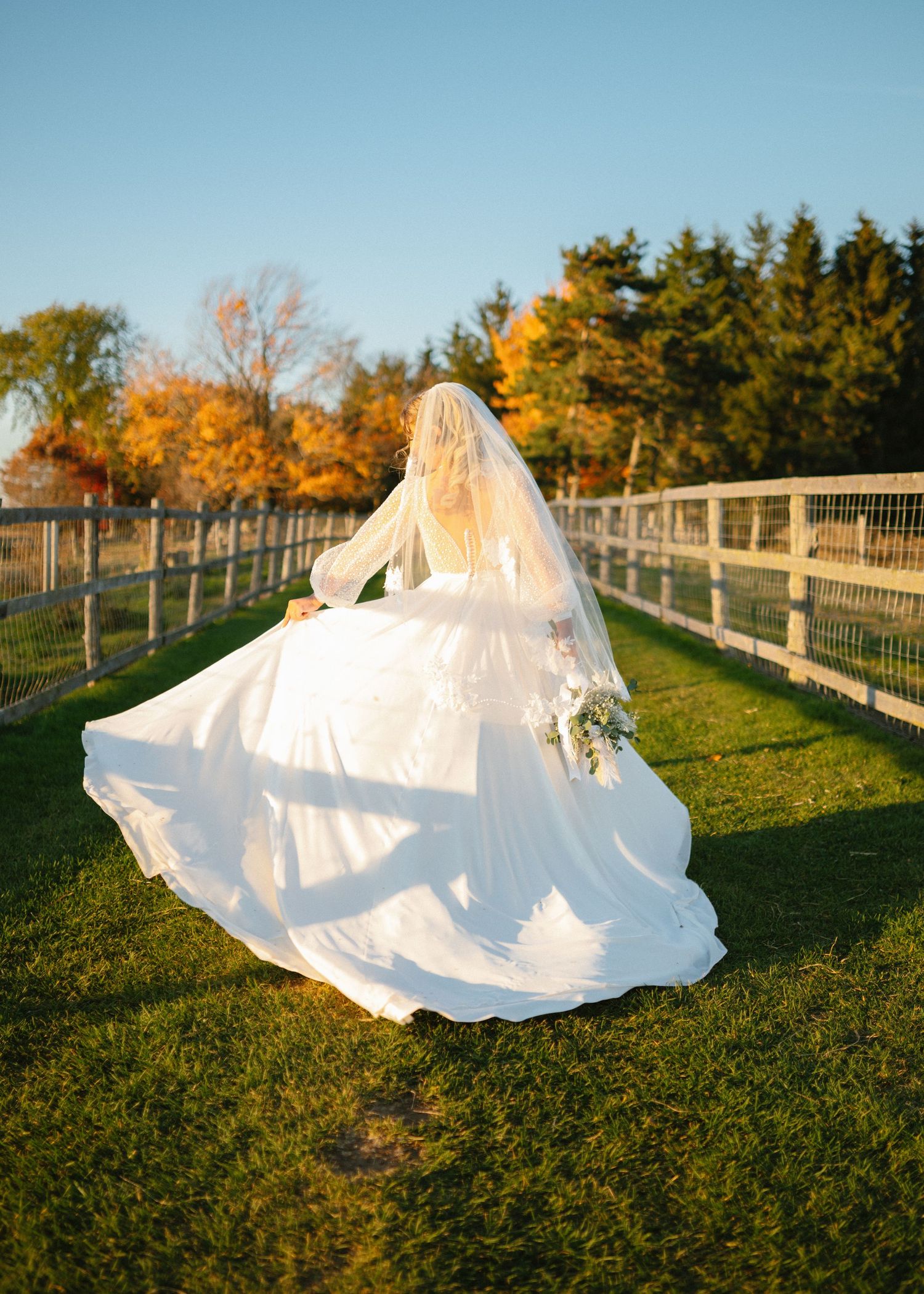 Bride in flowing white wedding gown and veil twirling on green grass with autumn trees in background.