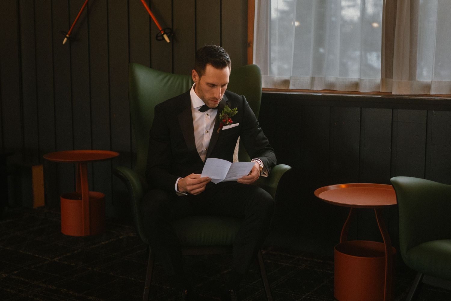 Groom in black tuxedo reads wedding vows letter while seated in green velvet chair in stylish venue.