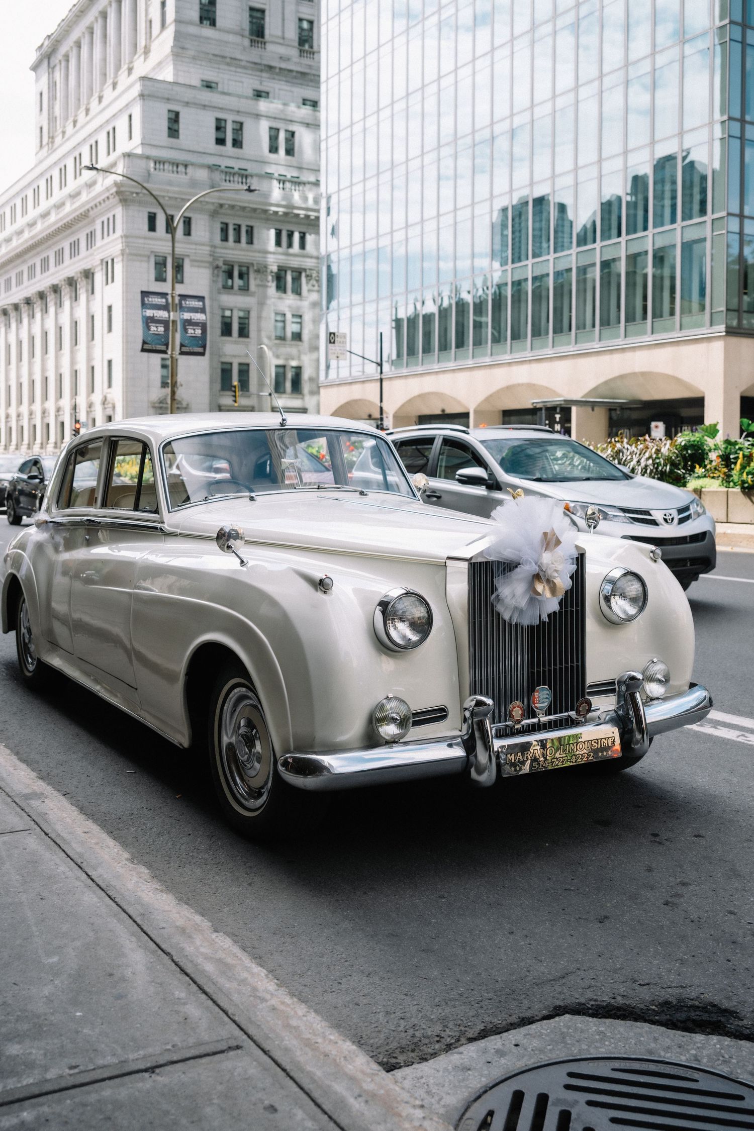 Vintage white Rolls-Royce decorated with wedding flowers driving through city streets.