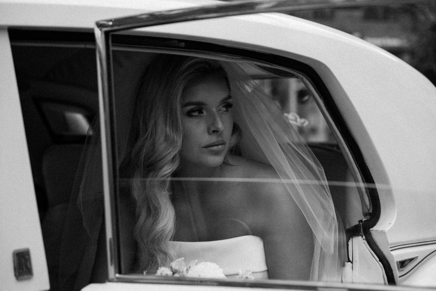 Black and white portrait of a bride in veil looking out of a vintage car window.