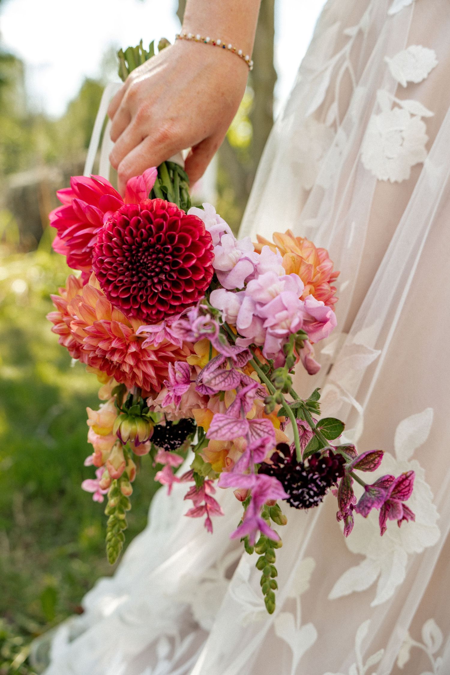 Bride holding a vibrant wedding bouquet with red dahlias and pink blooms in a floral lace gown.
