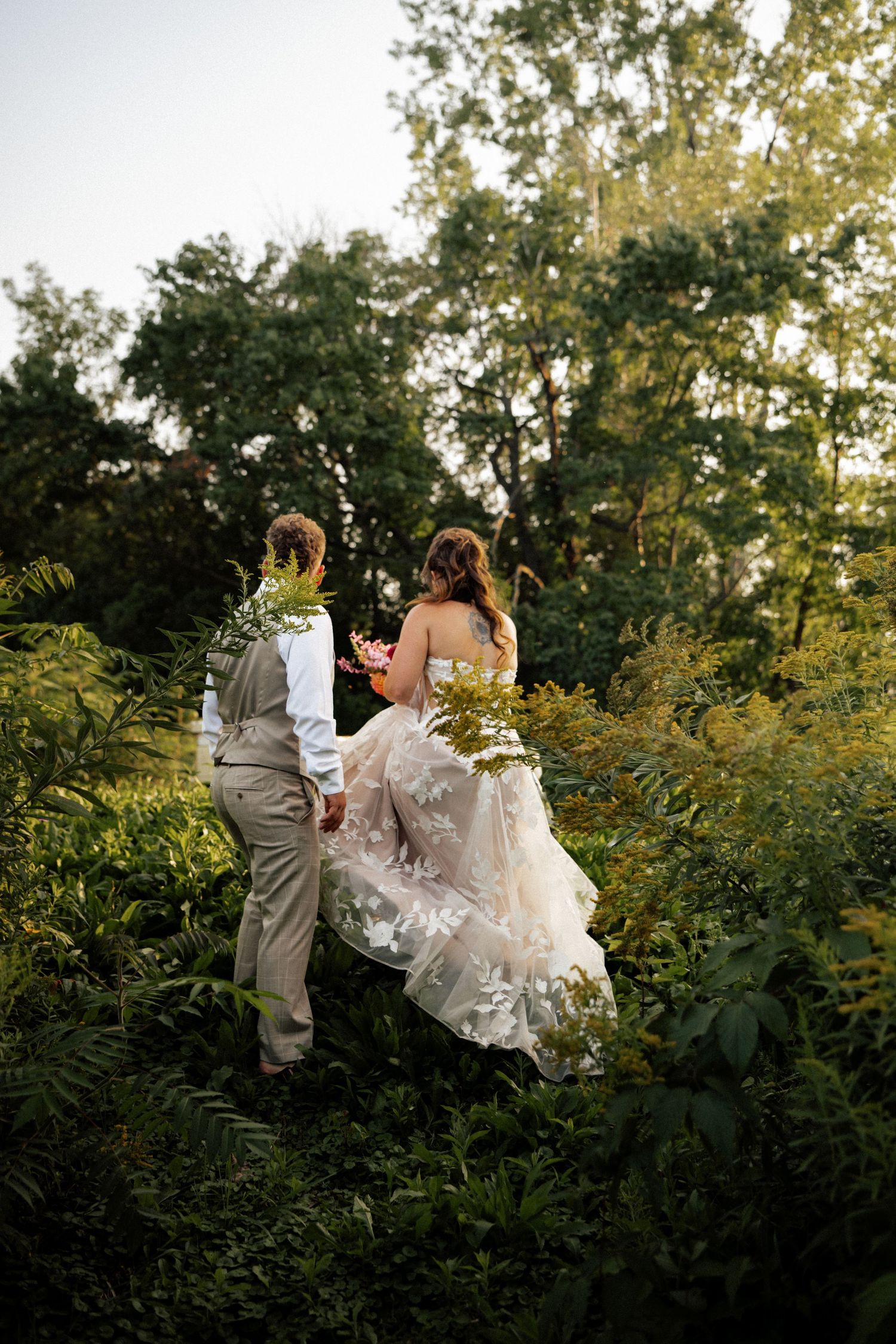 Bride in floral lace gown and groom walk hand in hand through lush green forest at golden hour.
