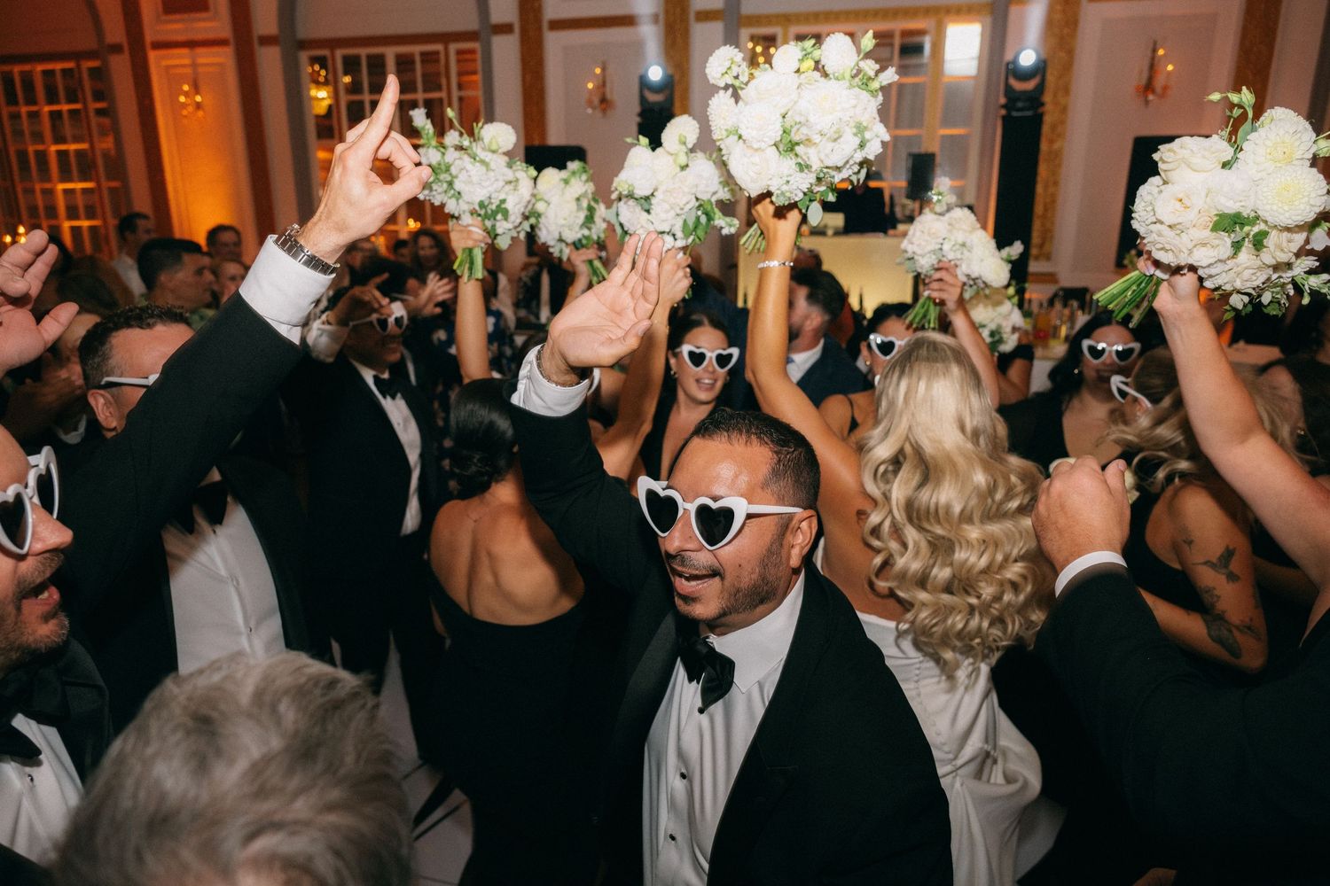 Wedding guests in tuxedos and gowns dance and celebrate while wearing heart-shaped white sunglasses.