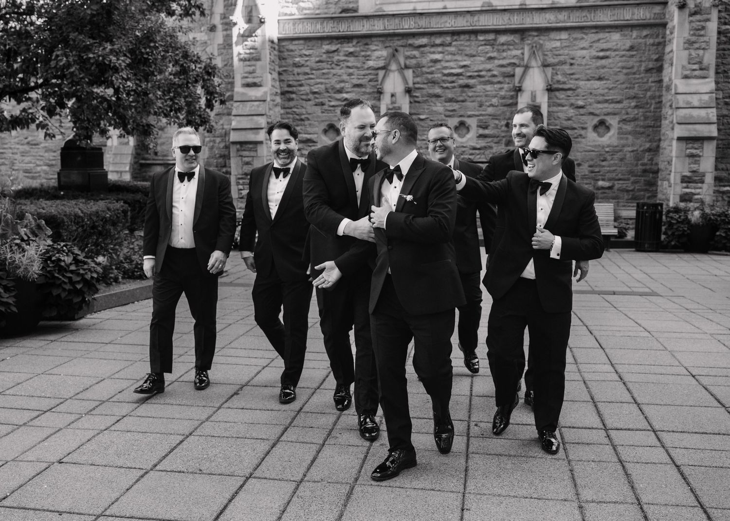 Black and white photo of a groom laughing with his groomsmen outside a stone church.