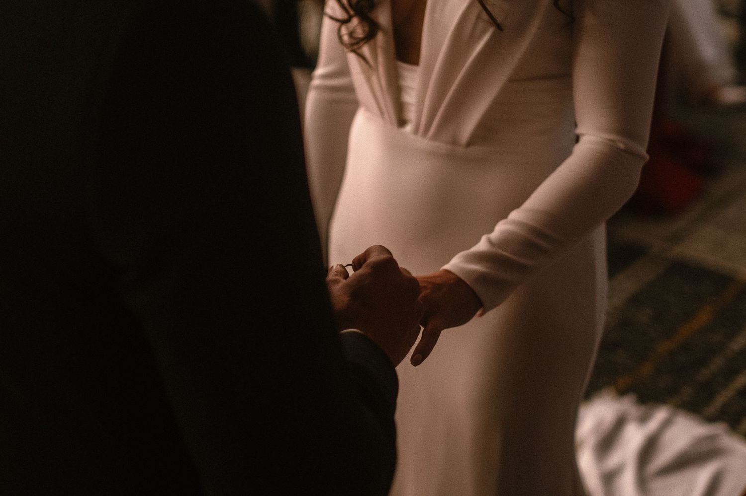 Couple holding hands in elegant wedding attire, bride in white long-sleeve gown in romantic dimly lit setting.