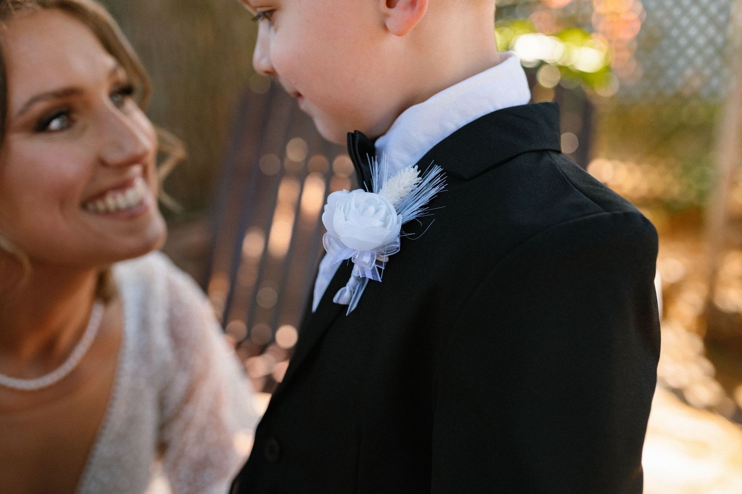 A smiling bride adjusts a boutonniere on a young boy in a black tuxedo at a wedding ceremony.