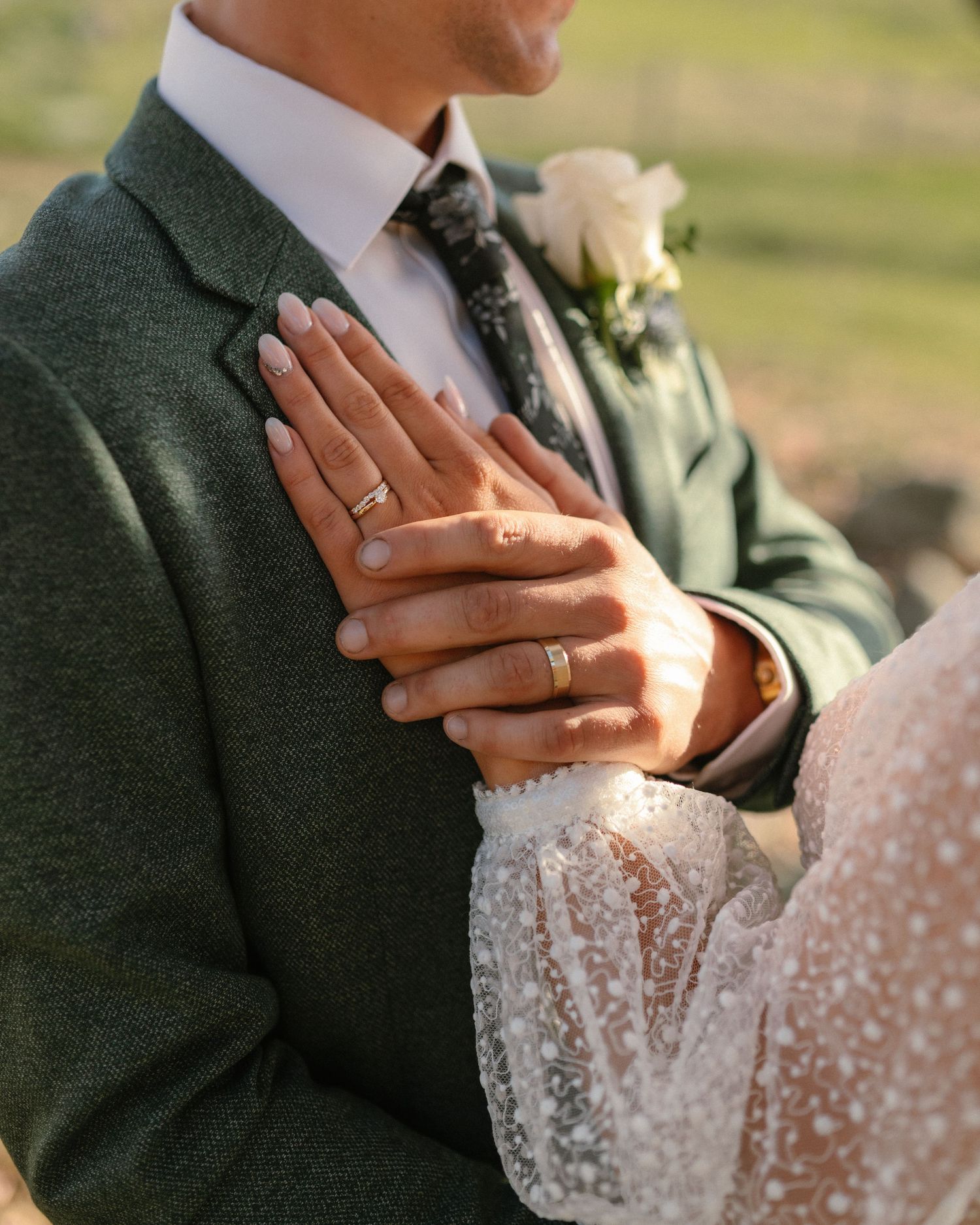 Close-up of bride and groom hands showing wedding rings, groom in green suit with white rose boutonniere.