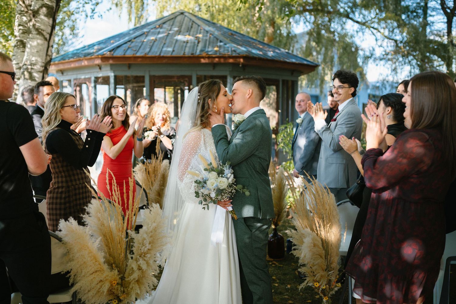 Bride and groom share first kiss at outdoor wedding ceremony while guests applaud near rustic gazebo.