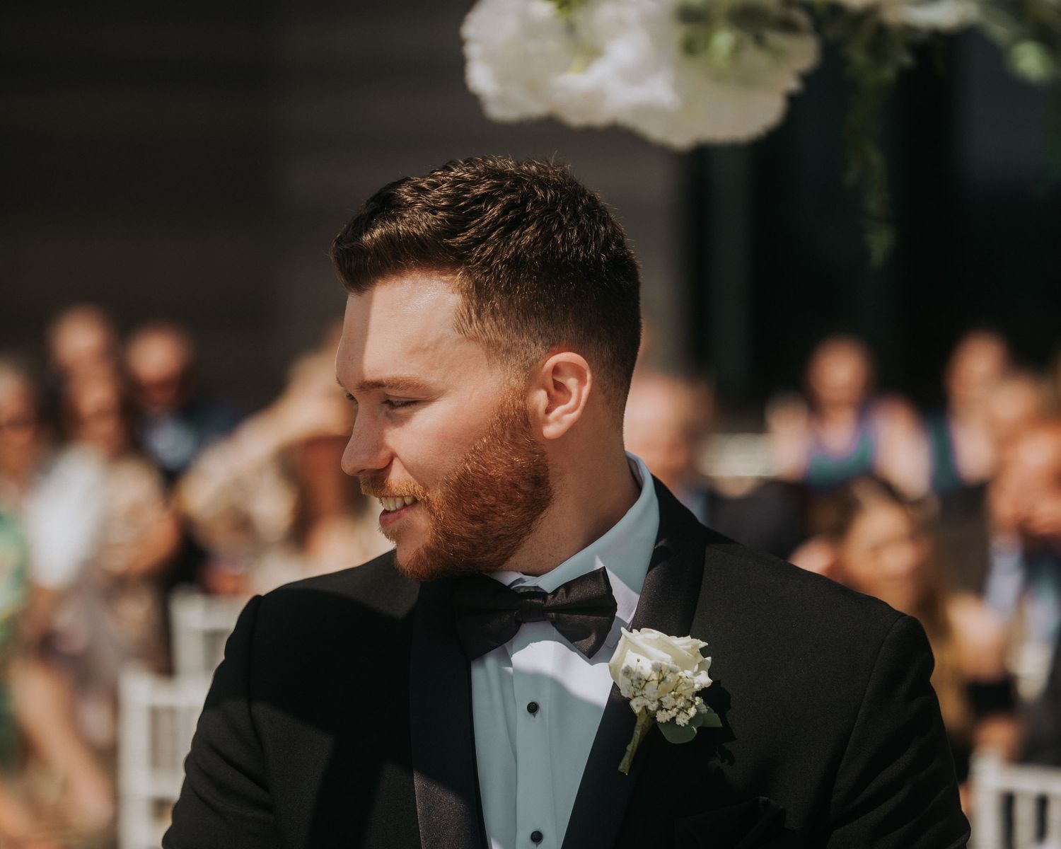 Handsome groom in black tuxedo with white boutonniere smiles at outdoor wedding ceremony.