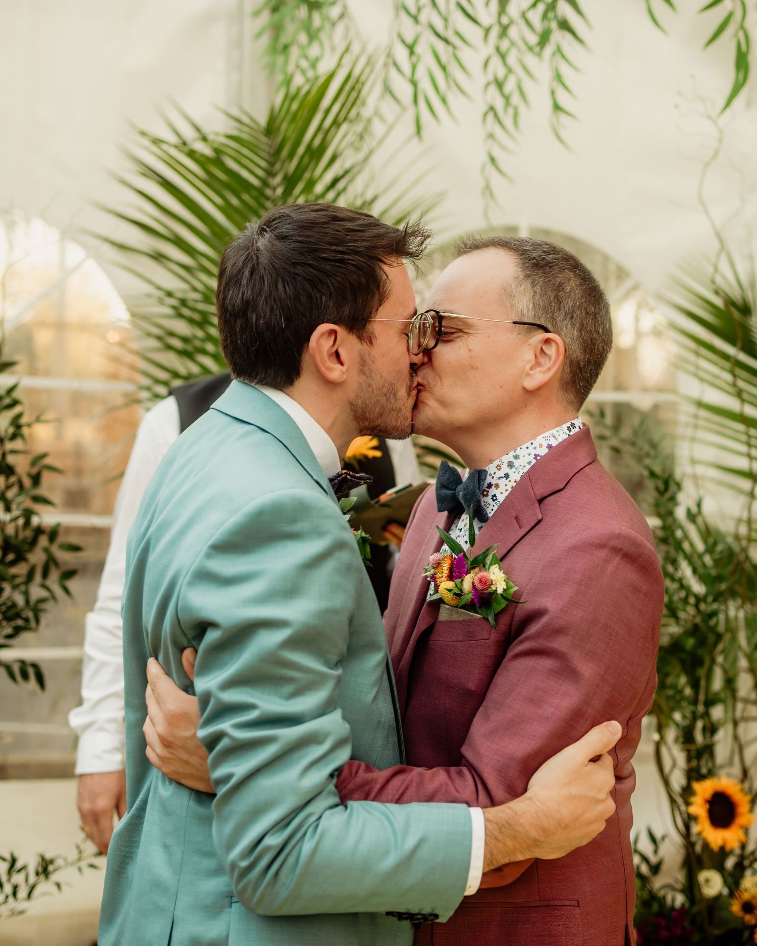Two grooms in colorful suits sharing a kiss at their wedding ceremony surrounded by tropical greenery.