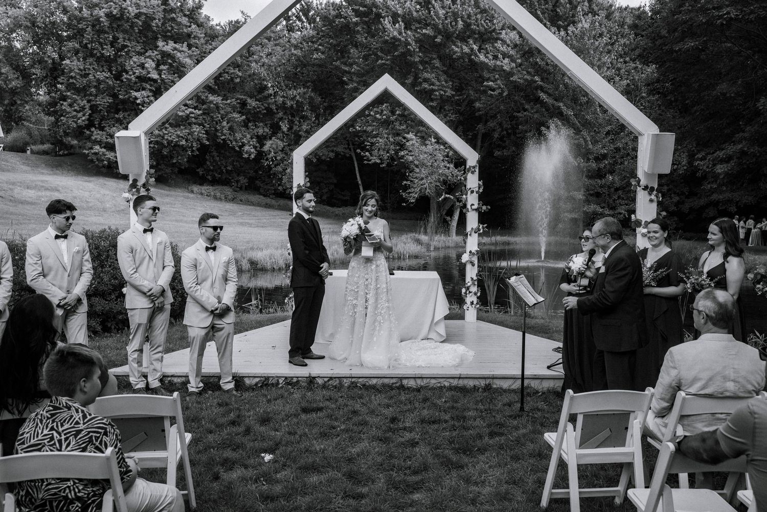 Black and white outdoor wedding ceremony with couple at floral arch, groomsmen, officiant, and guests on lawn near fountain.