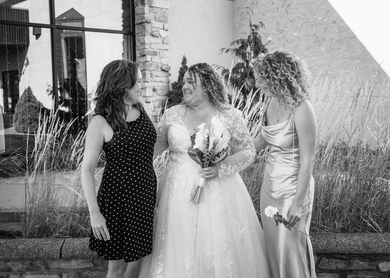 Black and white photo of a bride in a lace gown holding bouquet, flanked by two smiling women at an outdoor wedding.